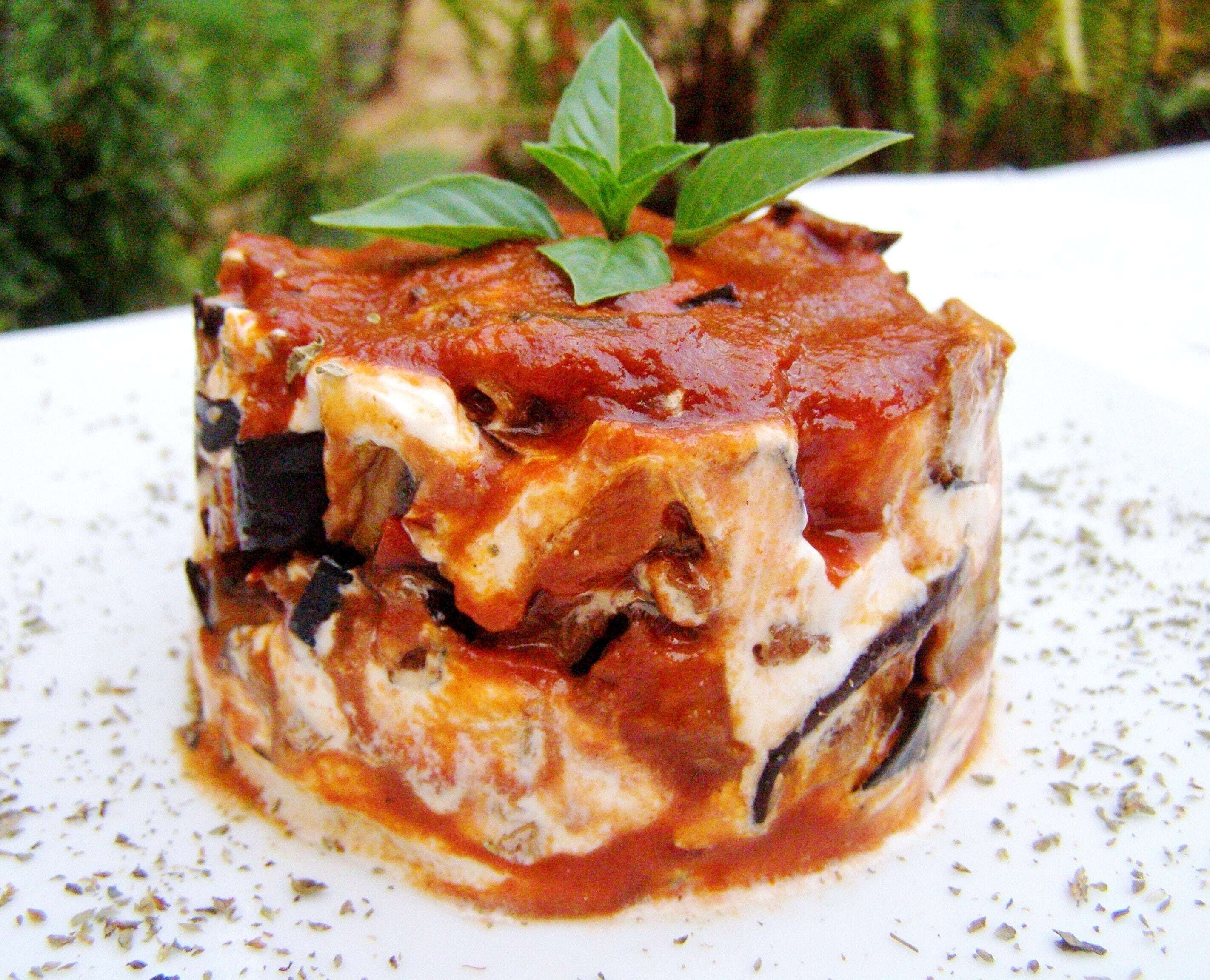 A close-up of a layered dish with a red sauce, possibly a type of lasagna, garnished with a basil leaf.