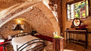 Un dormitorio rústico con paredes de piedra, una cama, una mesita de noche con espejo y una ventana con vistas a la vegetación.