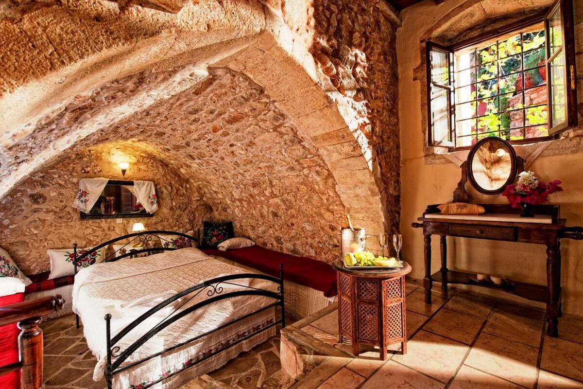 Une chambre rustique aux murs de pierre, avec un lit, une petite table surmontée d’un miroir et une fenêtre donnant sur un paysage verdoyant.