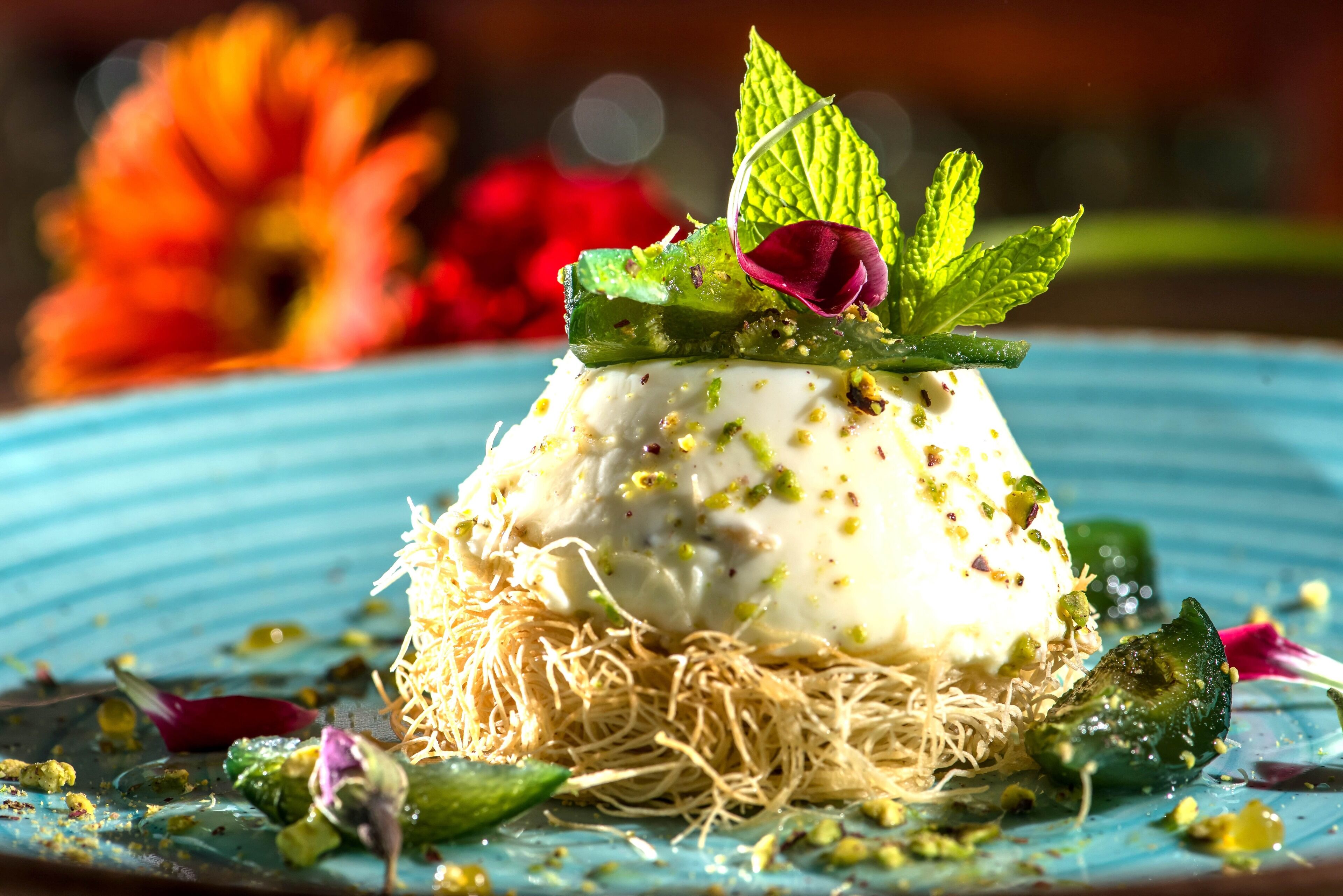 A dessert on a plate with a blue rim, garnished with mint leaves and a rose petal, surrounded by scattered nuts and a green leaf.