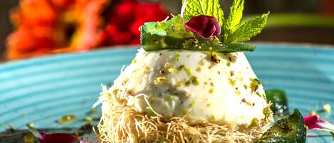 A dessert on a plate with a blue rim, garnished with mint leaves and a rose petal, surrounded by scattered nuts and a green leaf.