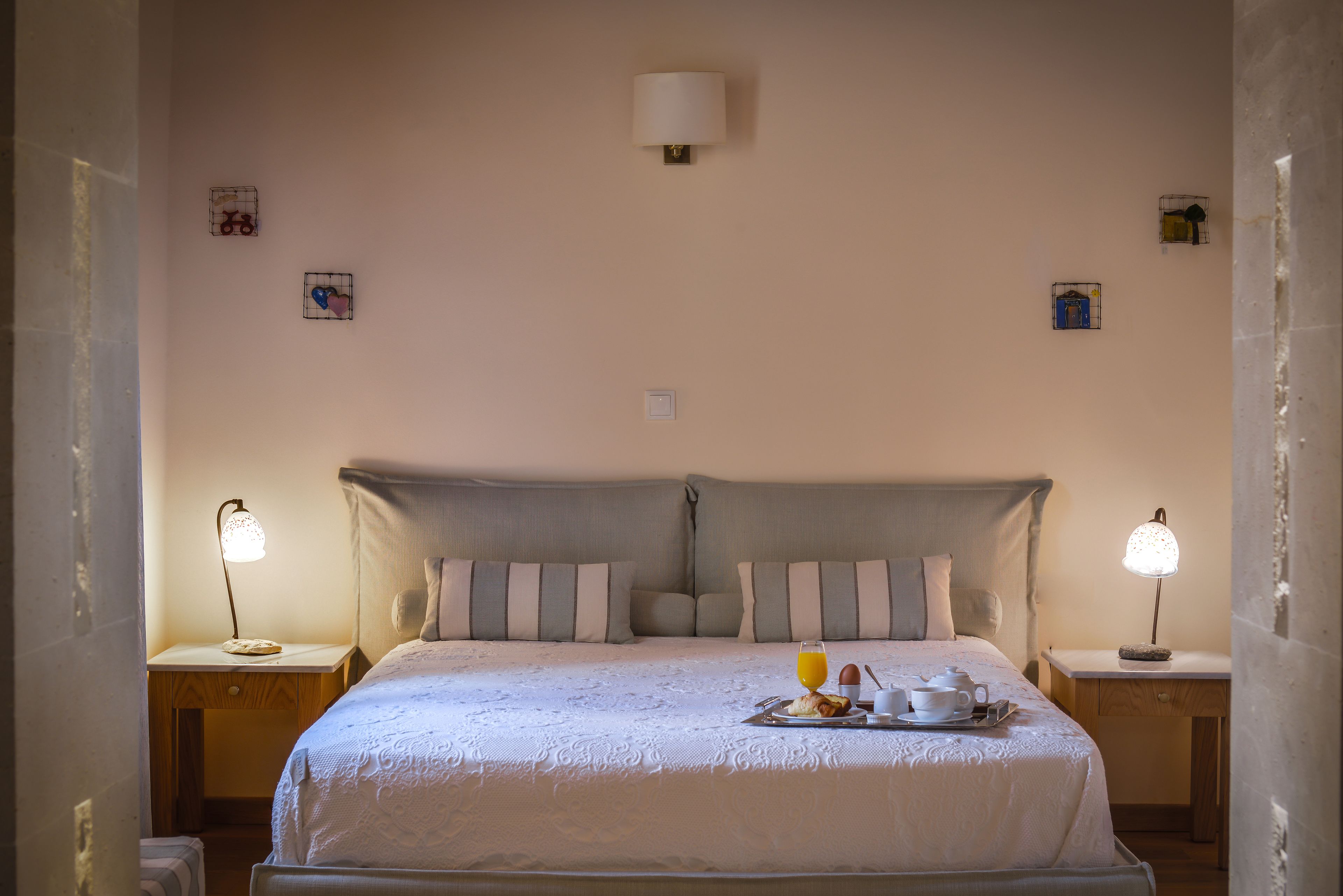 Una cama bien hecha con cubrecama blanco, dos lámparas de noche, una mesita con artículos para el desayuno y una lámpara de pared.