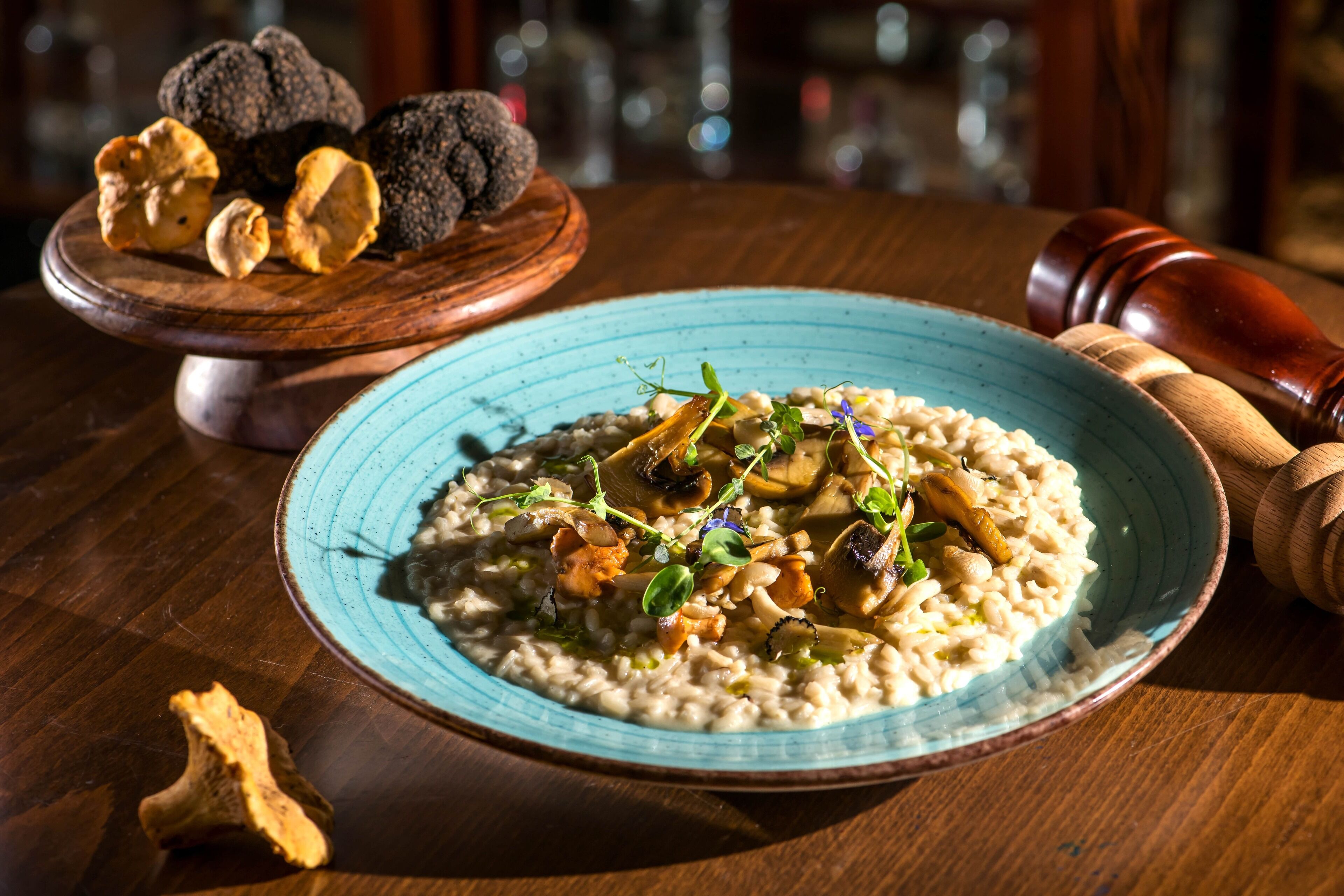A plate of risotto with mushrooms and herbs, a wooden board with truffles, and a pepper grinder on a wooden table.