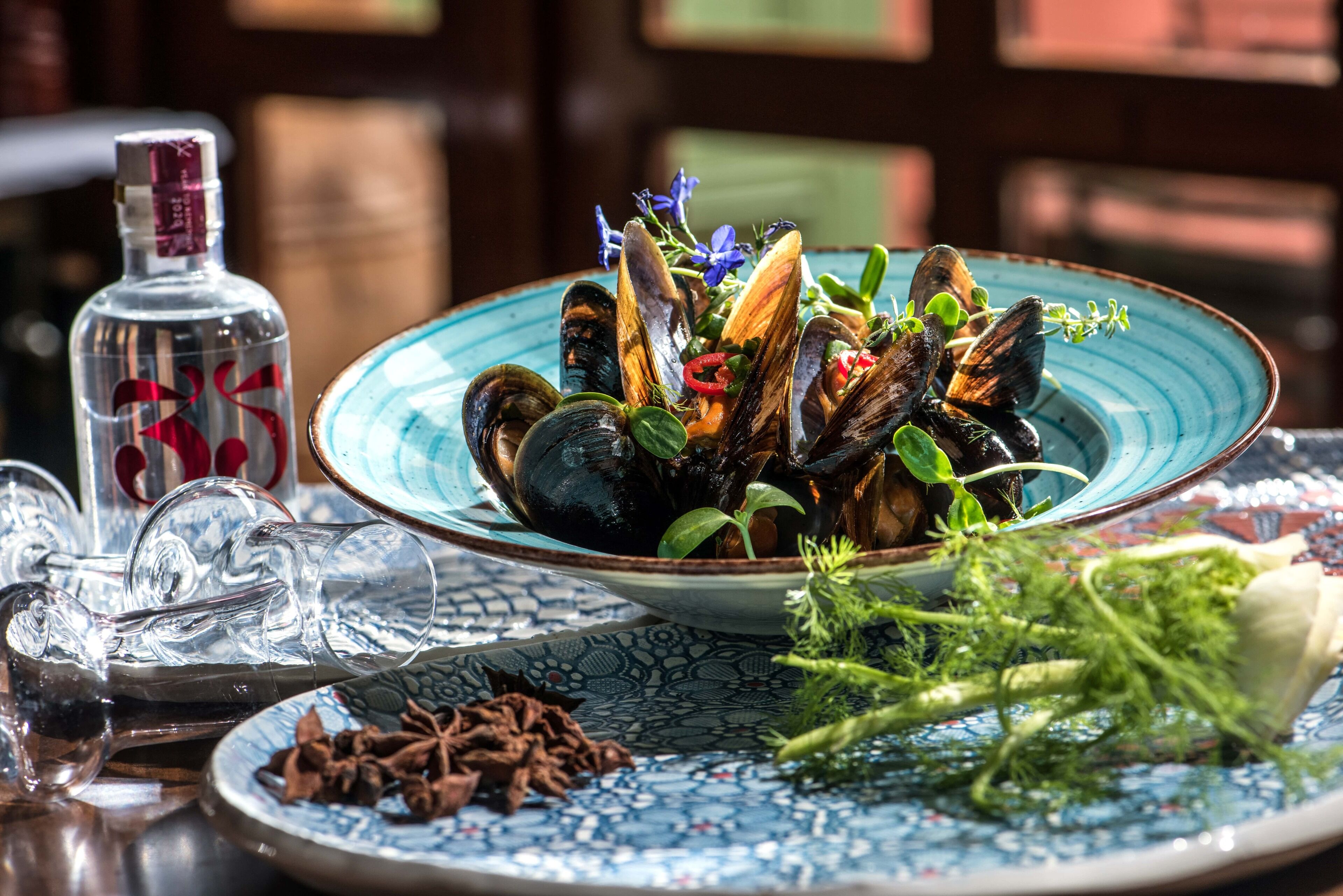 Une table avec une assiette bleue remplie de moules, décorée d’herbes et de fleurs, une bouteille en verre et une assiette contenant des étoiles de badiane.