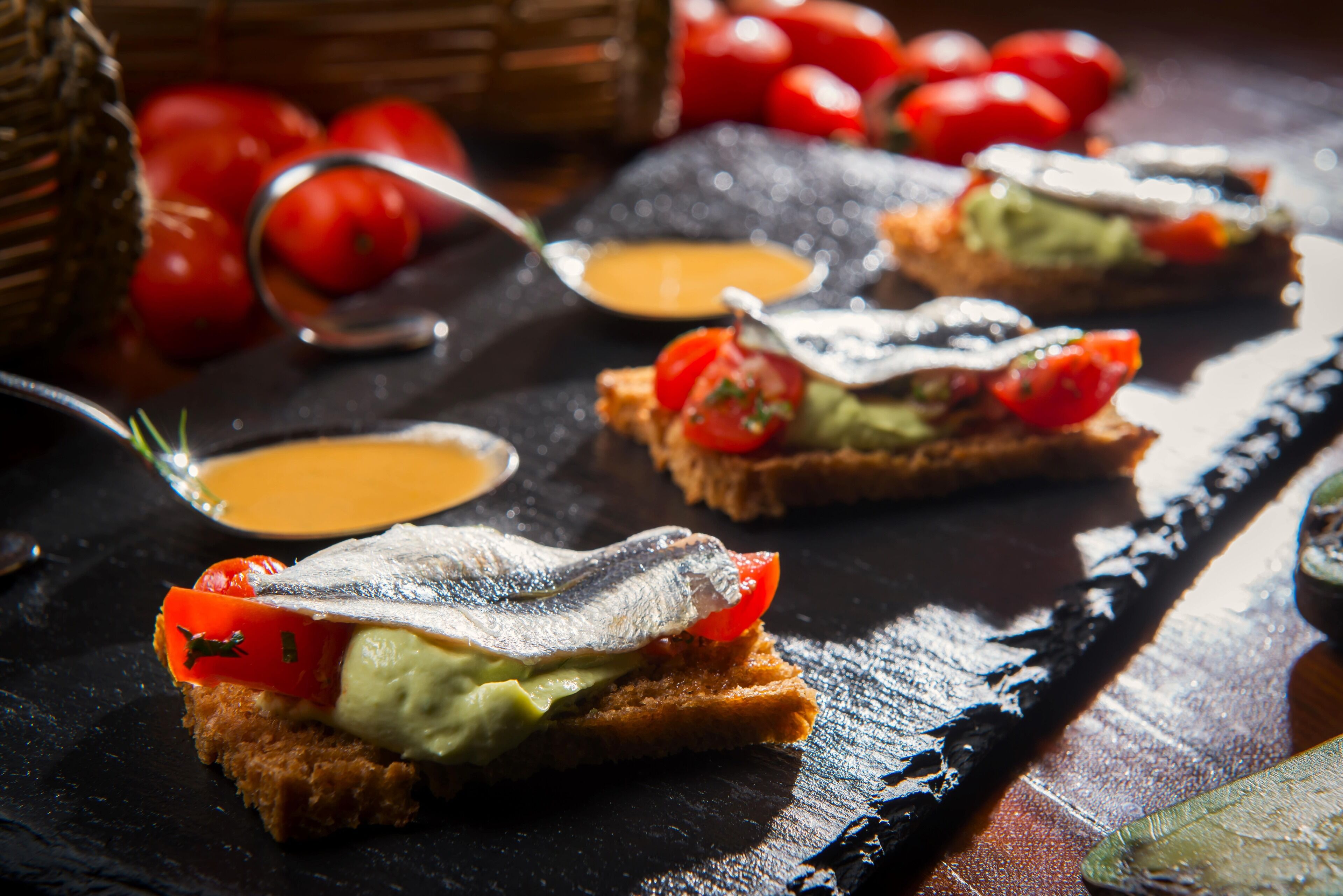 Close-up van een bord met vis, avocado en tomaat op een sneetje brood, met een lepel en een klein schaaltje saus erbij.