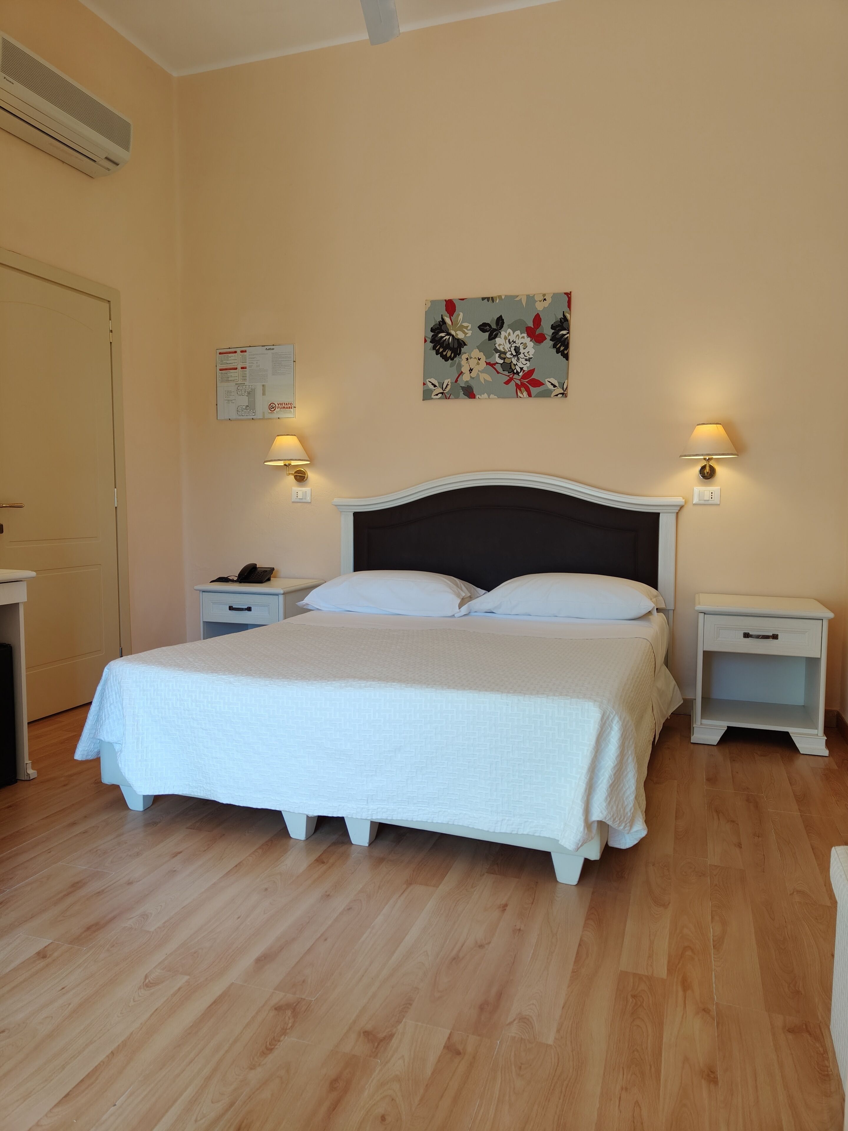 Un dormitorio con una cama, dos mesitas de noche, aire acondicionado instalado en la pared, una obra de arte floral y una lámpara fijada a la pared.