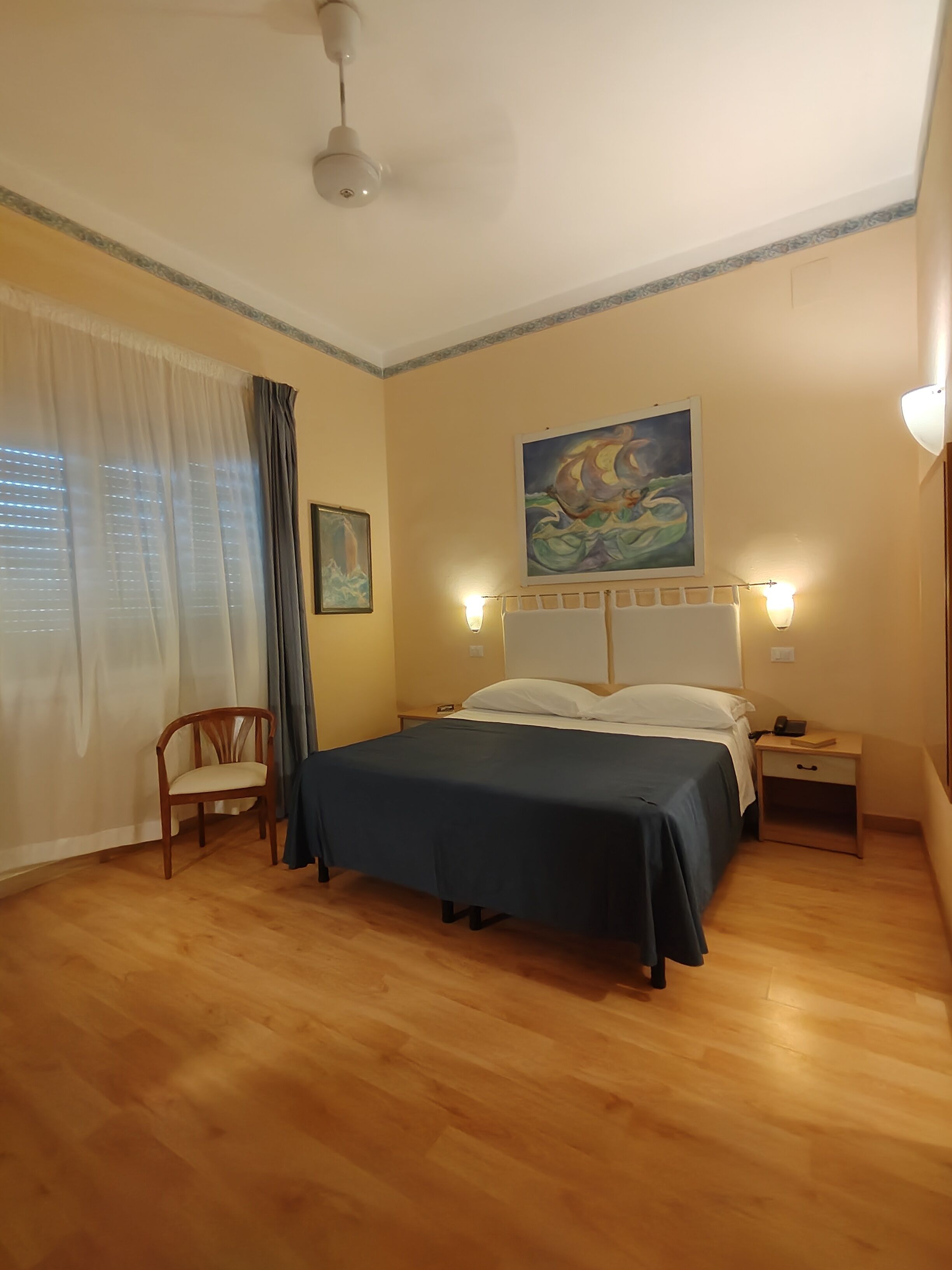 A bedroom with a bed, a chair, a nightstand, a painting, and a ceiling fan.