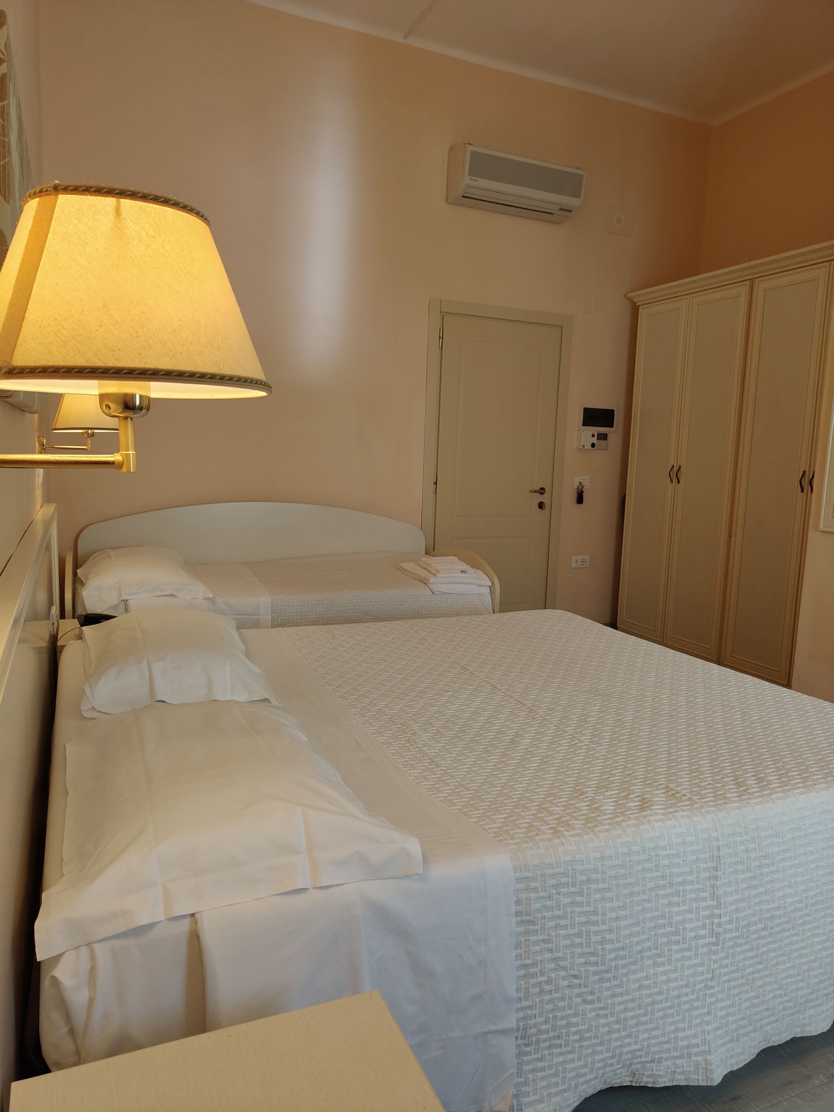 A hotel room with a bed, a bedside lamp, an air conditioning unit, a wardrobe, and a door.