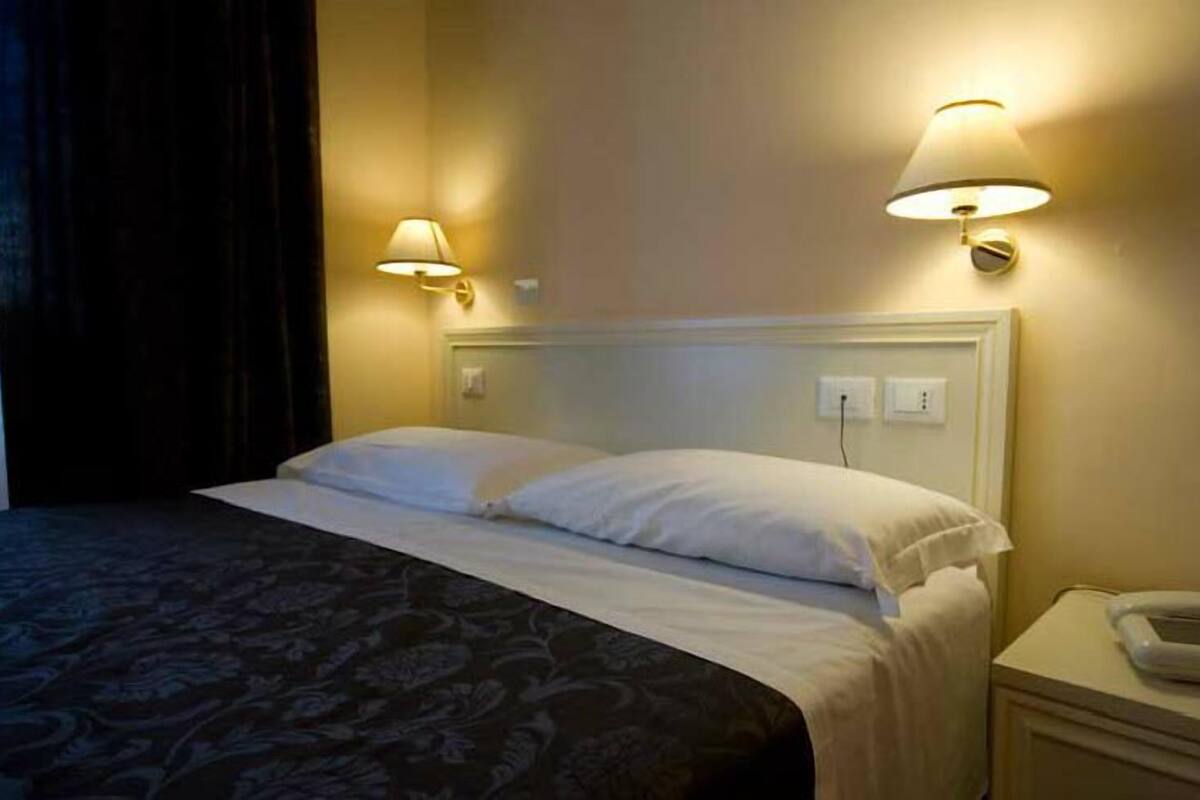 A hotel room with a bed, two wall-mounted lamps, a bedside phone, and a power outlet.