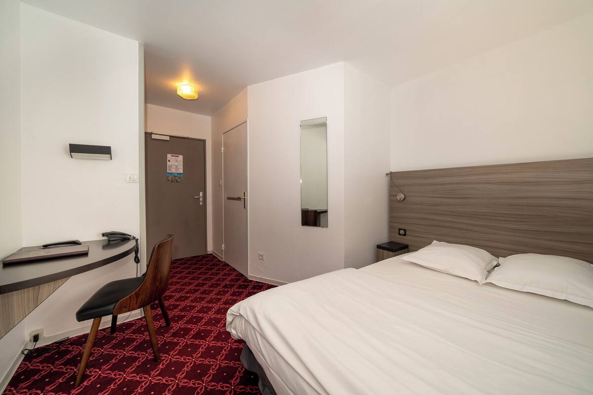 Une chambre d’hôtel avec un lit, un bureau, une chaise, un miroir et un tapis rouge à motifs.