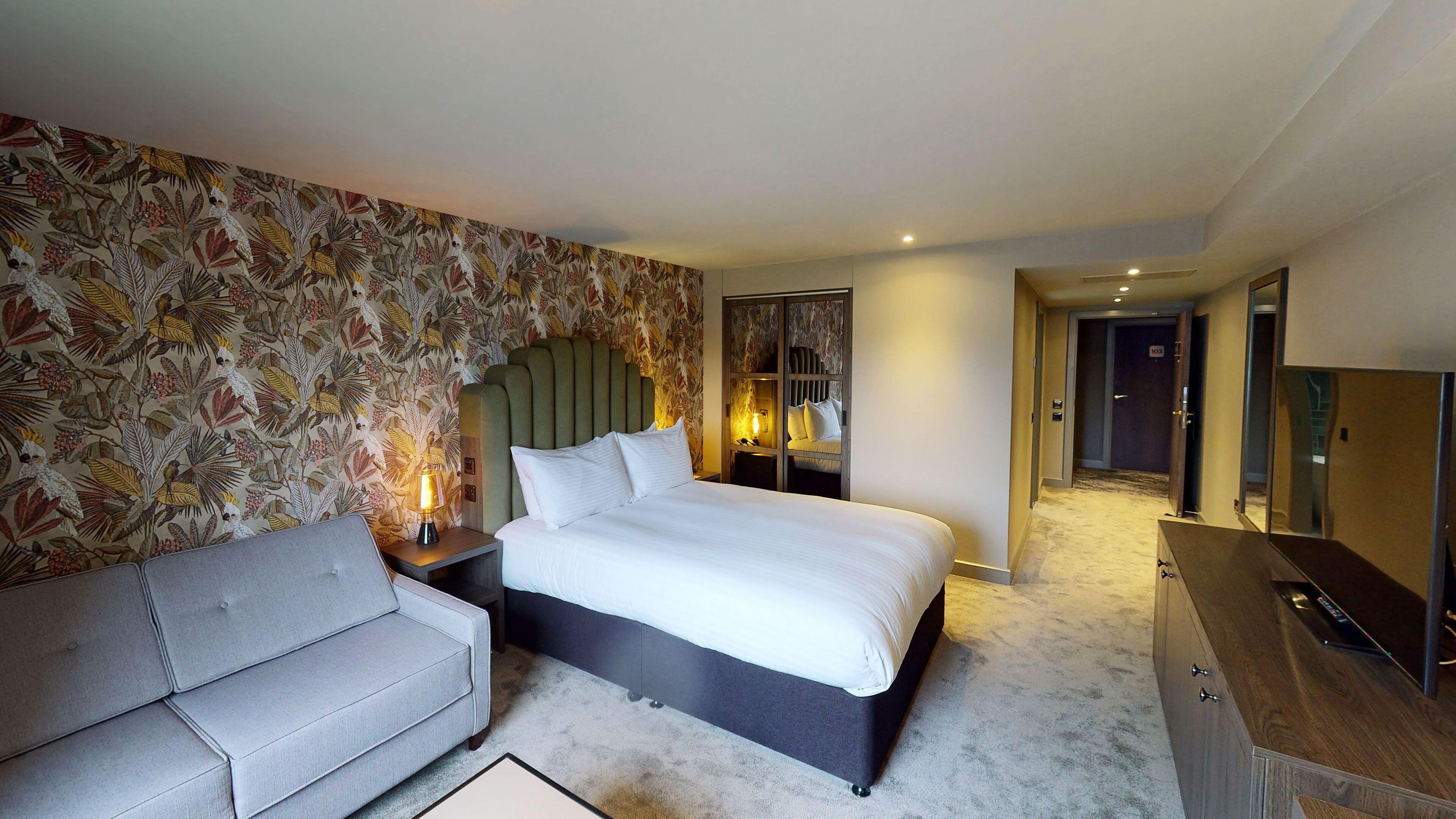 A hotel room with a bed, a nightstand with a lamp, a sofa, a wardrobe, and a mirror.