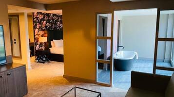 A modern hotel room with a sofa, a glass coffee table, a TV, a floral-patterned headboard, and a view of a bathroom with a bathtub.