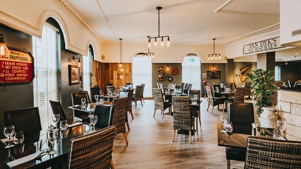 Een restaurantinterieur met meerdere eettafels en stoelen, houten lambrisering aan de wanden, en een bord met de tekst 'Van Dyk's Nurseries'.