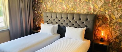 Habitación de hotel con dos camas, cabecero acolchado, pared con diseño, mesita de noche con lámpara y ventana con cortinas.