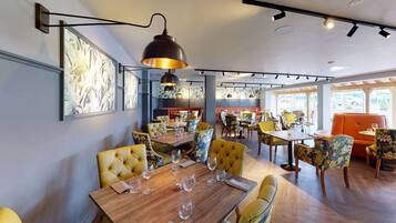 A restaurant interior with wooden tables, yellow upholstered chairs, and a large pendant light.