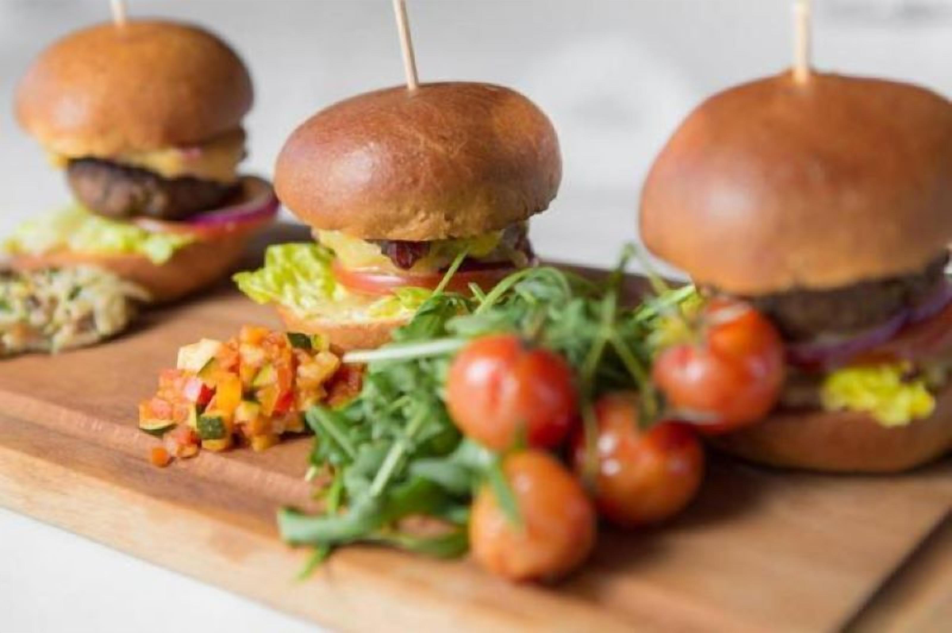 Three burgers with buns on a wooden board, accompanied by a side of mixed vegetables and cherry tomatoes.