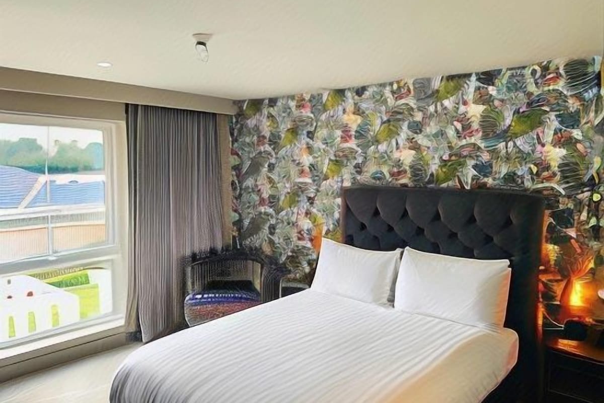 A hotel room with a large bed, a headboard with a floral pattern, a window with a view of a building, and a nightstand with a lamp.
