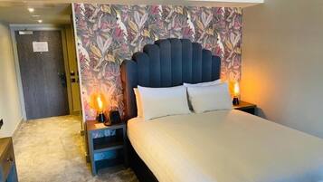 Une chambre d’hôtel avec un grand lit, deux tables de chevet avec des lampes, une tête de lit à motifs floraux et une porte donnant sur une autre pièce.