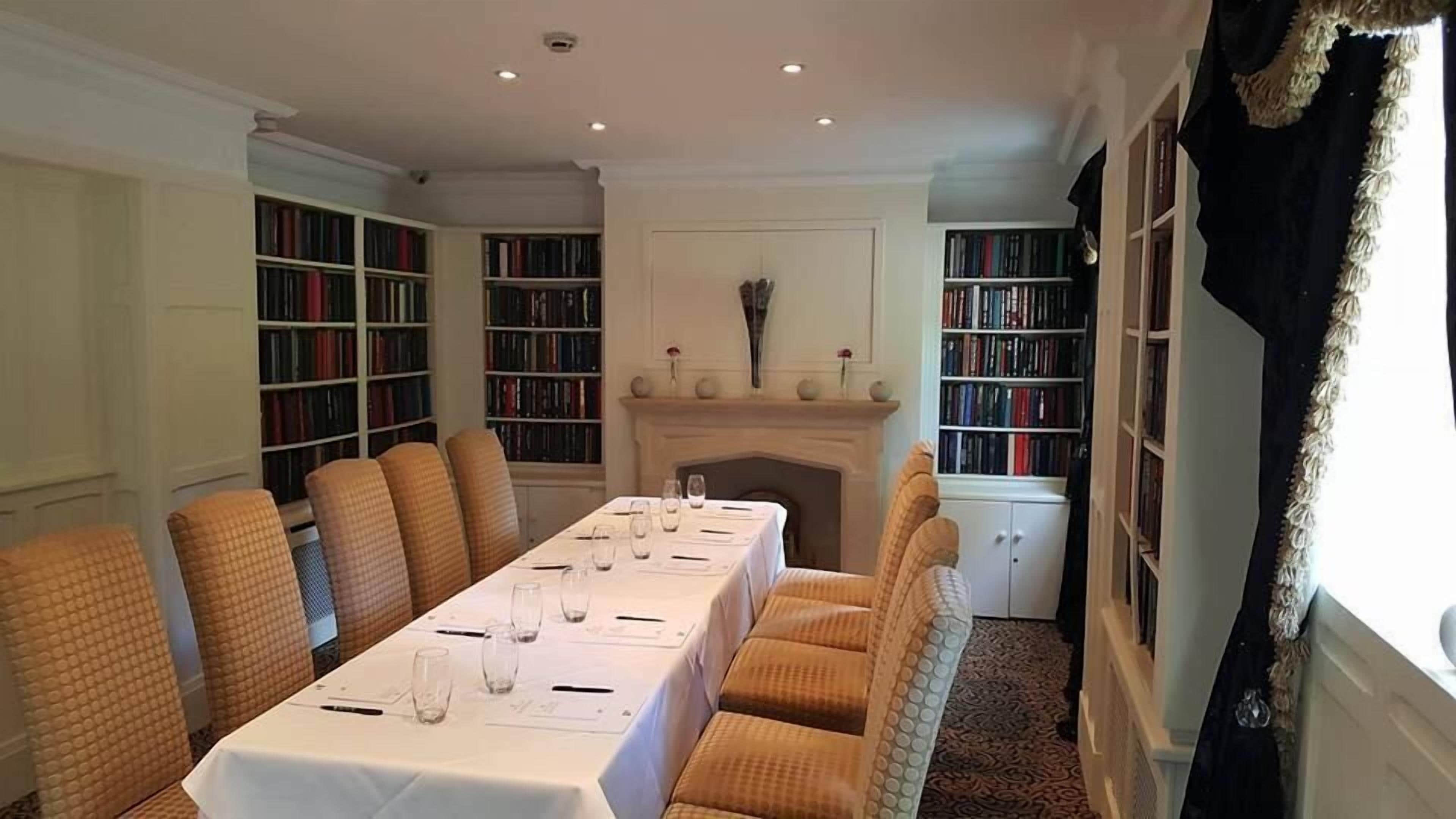 Un comedor formal con una larga mesa dispuesta para seis personas, flanqueada por estanterías, una chimenea y un ventanal con cortinas.