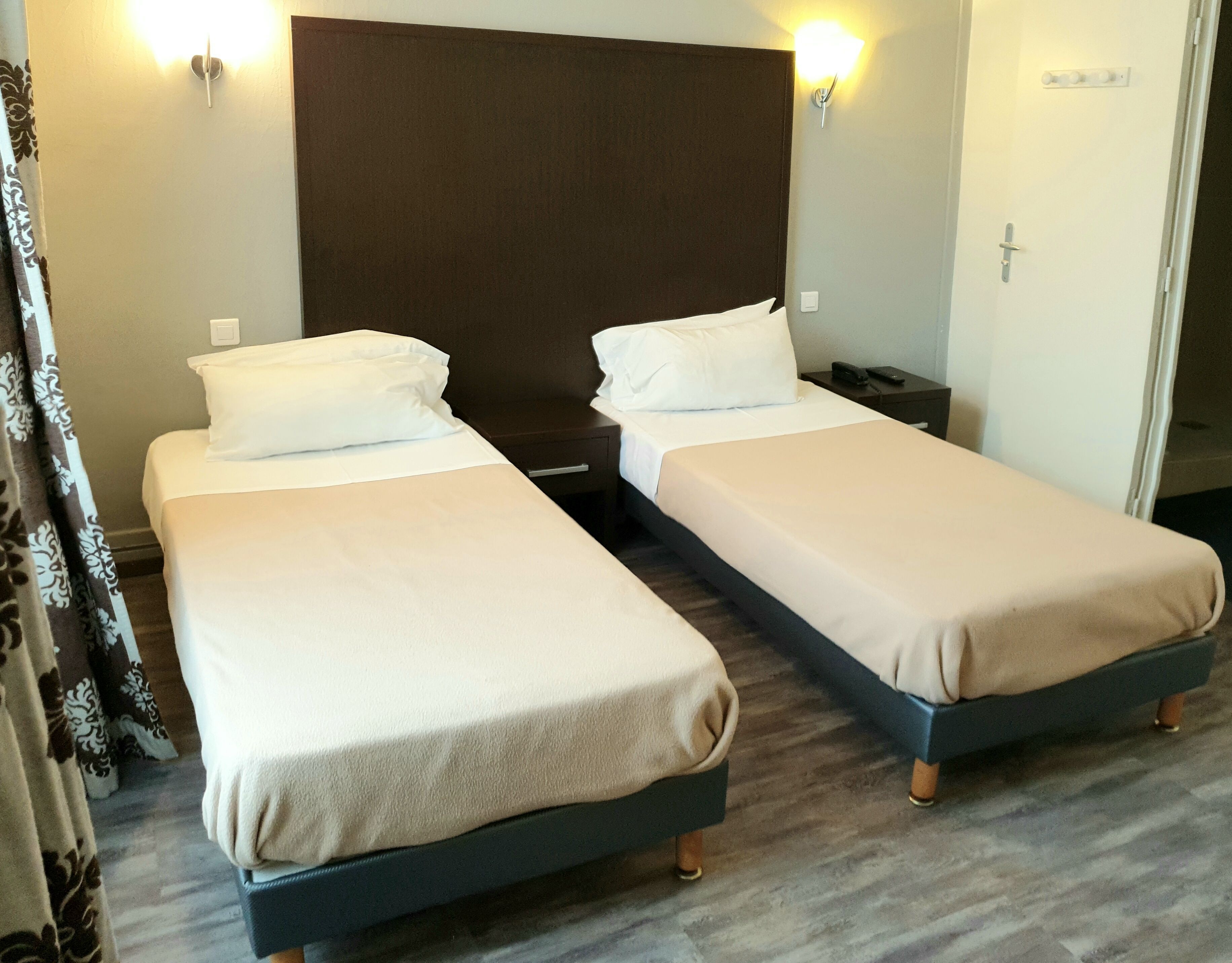 A hotel room with two single beds, each with a white pillow and a beige blanket. There is a dark headboard behind the beds, and a small bedside table with a remote control and a phone.