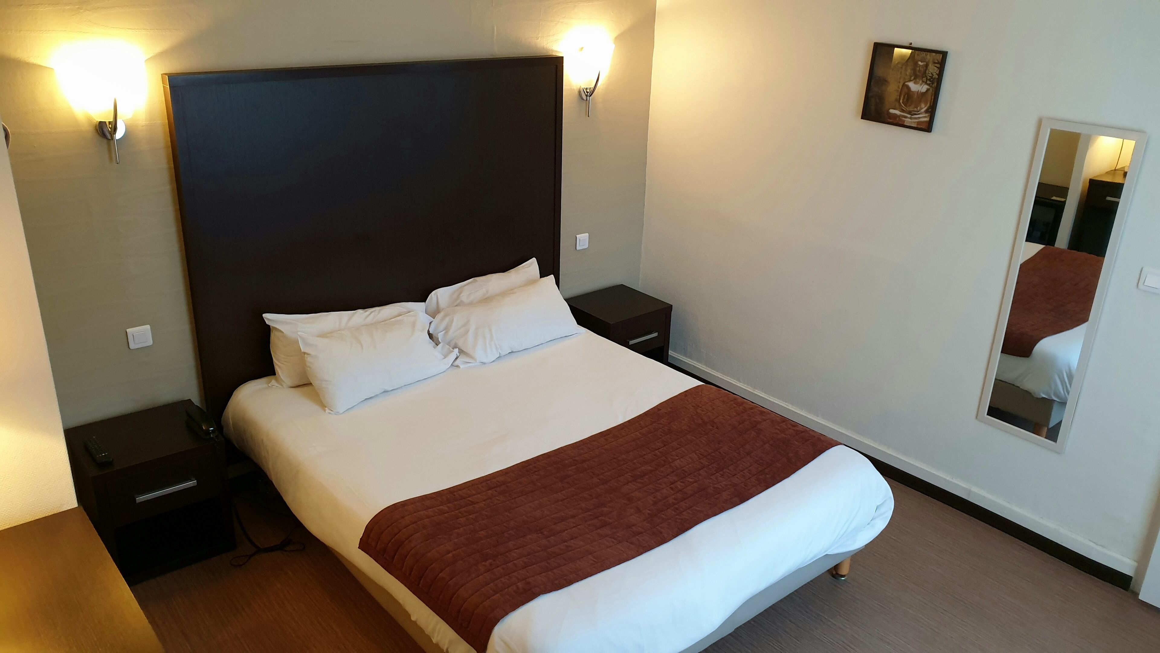 A hotel room with a large bed, two bedside tables, a nightstand, a wall-mounted light, a small painting, and a mirror.