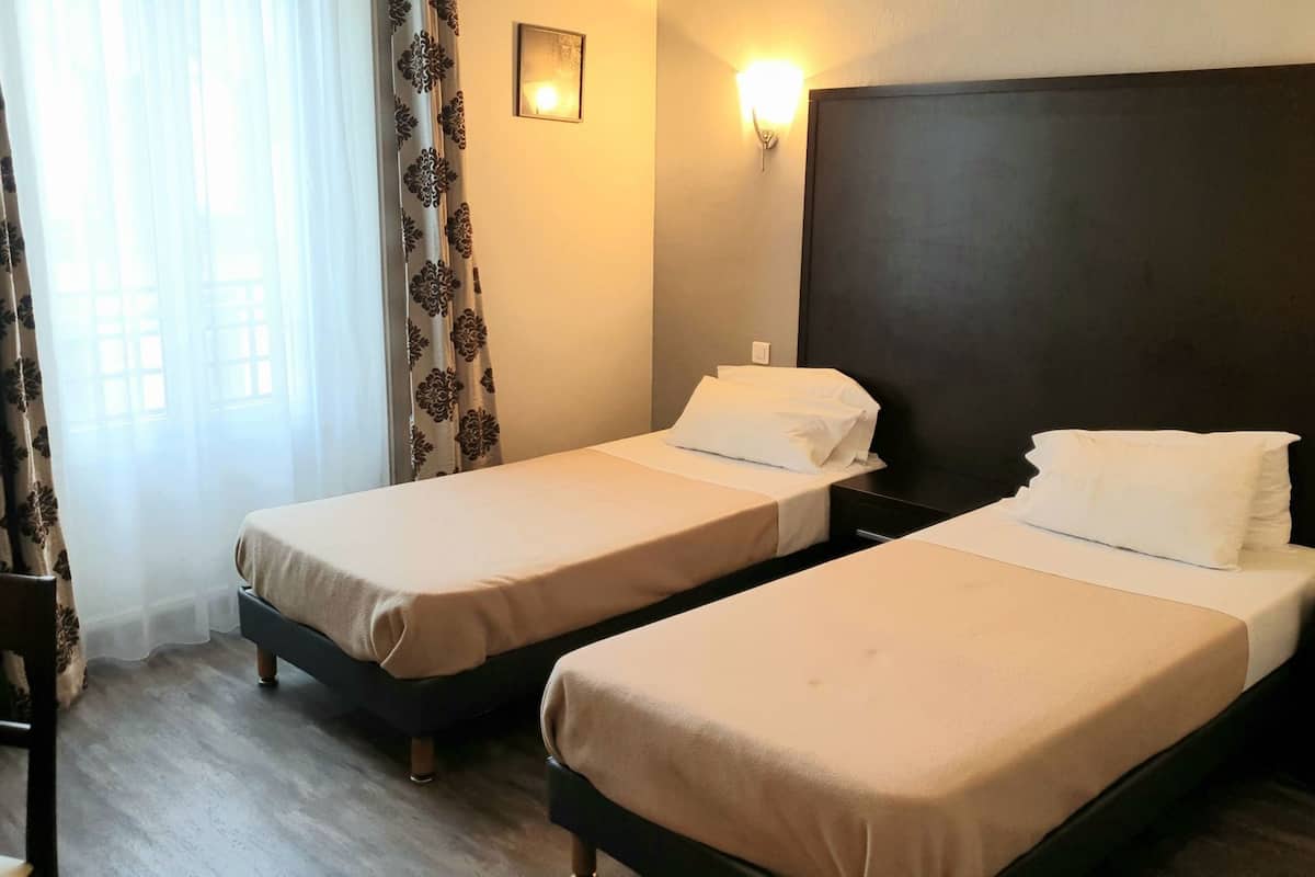 Une chambre d’hôtel avec deux lits simples, un bureau, une chaise, une lampe fixée au mur et une fenêtre avec des rideaux.