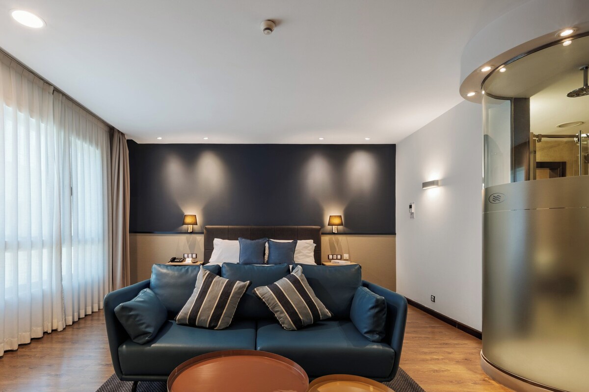 Una habitación de hotel moderna con un sofá azul oscuro, dos lámparas, una mesita de centro redonda y un espejo grande.