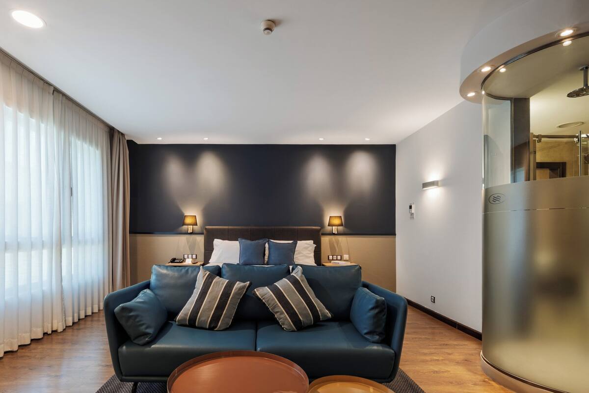 Una habitación de hotel moderna con un sofá azul oscuro, dos lámparas, una mesita de centro redonda y un espejo grande.