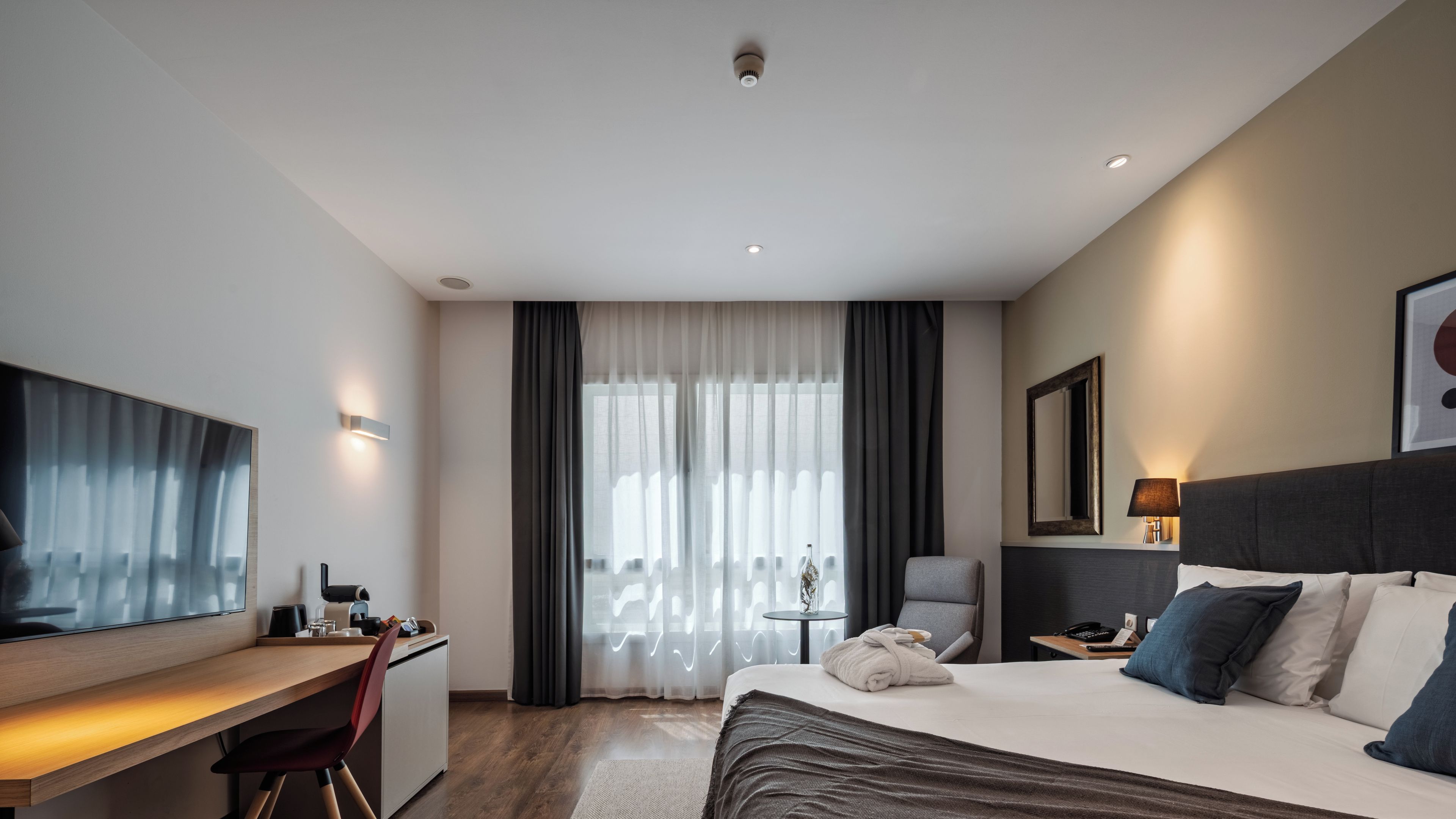Et moderne hotelværelse med seng, skrivebord, stol og et stort vindue med gardiner.