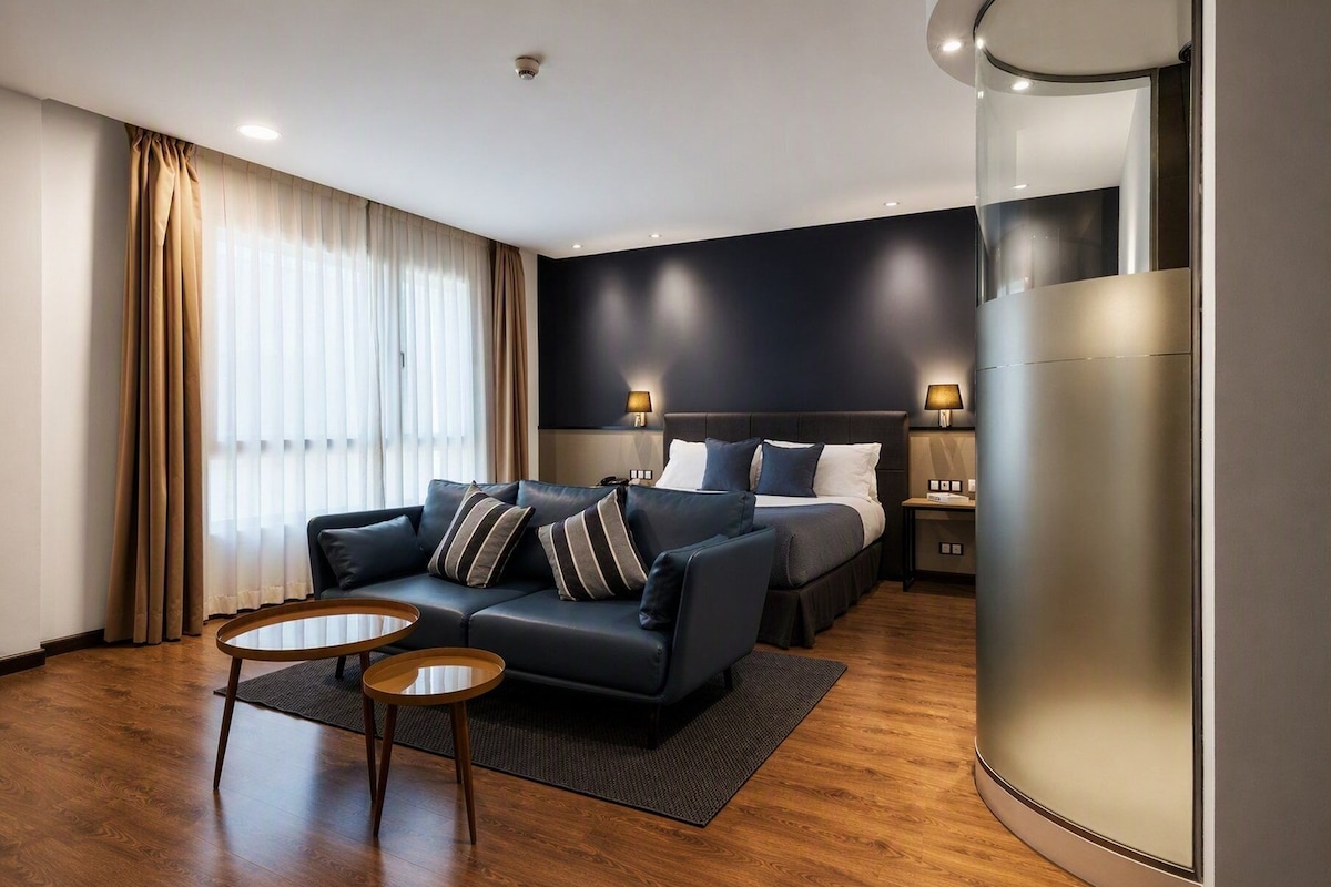 A modern hotel room with a dark blue headboard bed, a leather sofa, two small wooden tables, and a large cylindrical column.