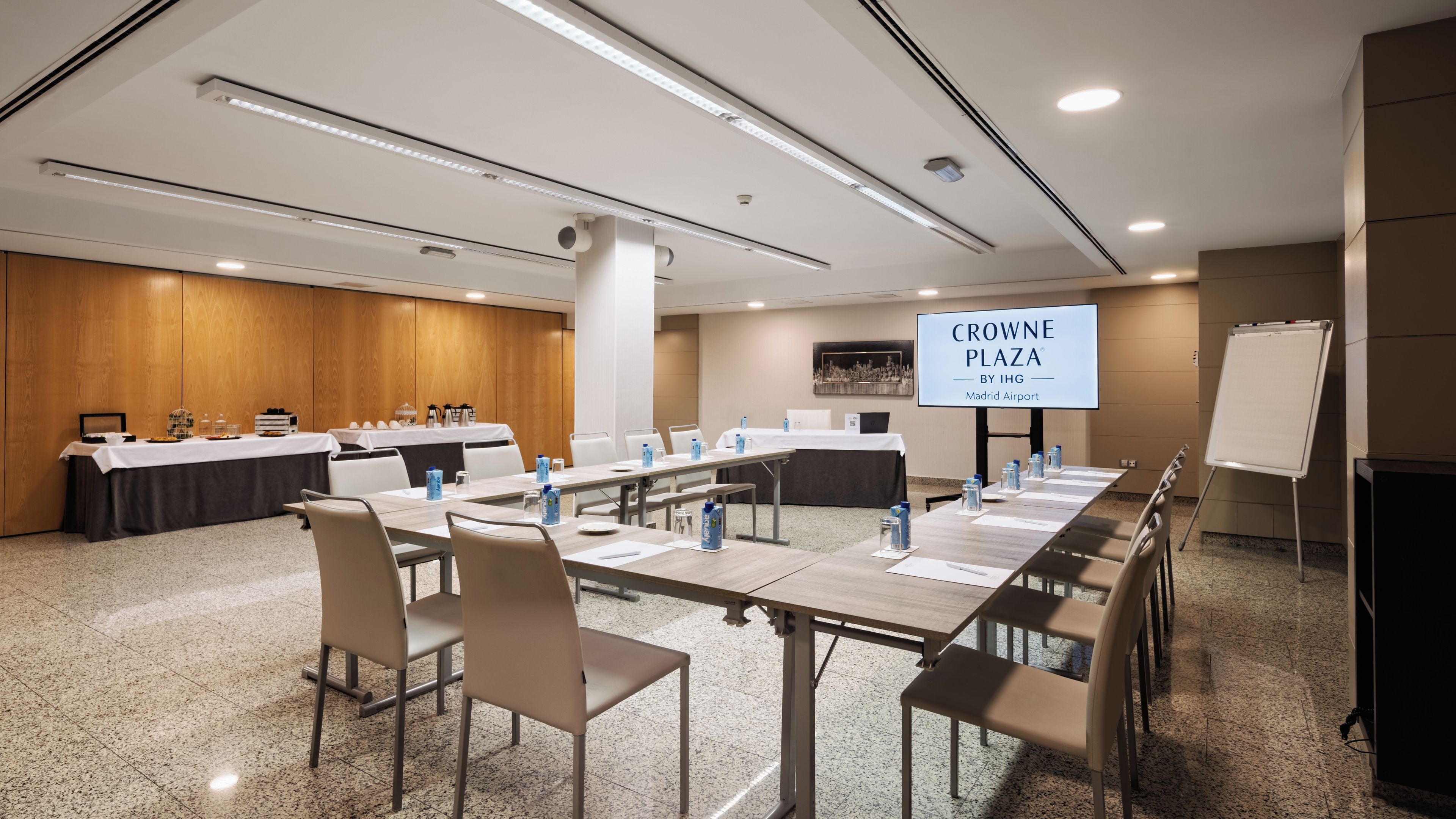 A conference room with a long table, chairs, a whiteboard, and a screen displaying 'CROWNE PLAZA BY IHG Madrid Airport'.
