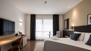 Habitació d'hotel amb llit, escriptori, cadira i una finestra gran amb cortines.