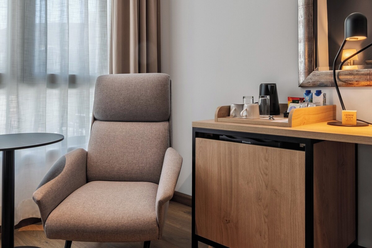 Una habitación de hotel moderna con un sillón gris, una pequeña mesa redonda, un escritorio de madera con una lámpara y una ventana con cortinas translúcidas.