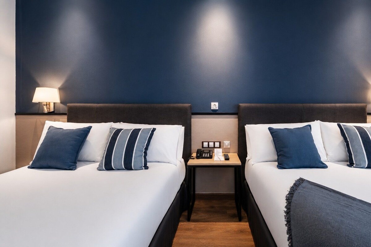 Quarto de hotel com duas camas, uma mesinha de cabeceira e luminárias fixadas na parede.