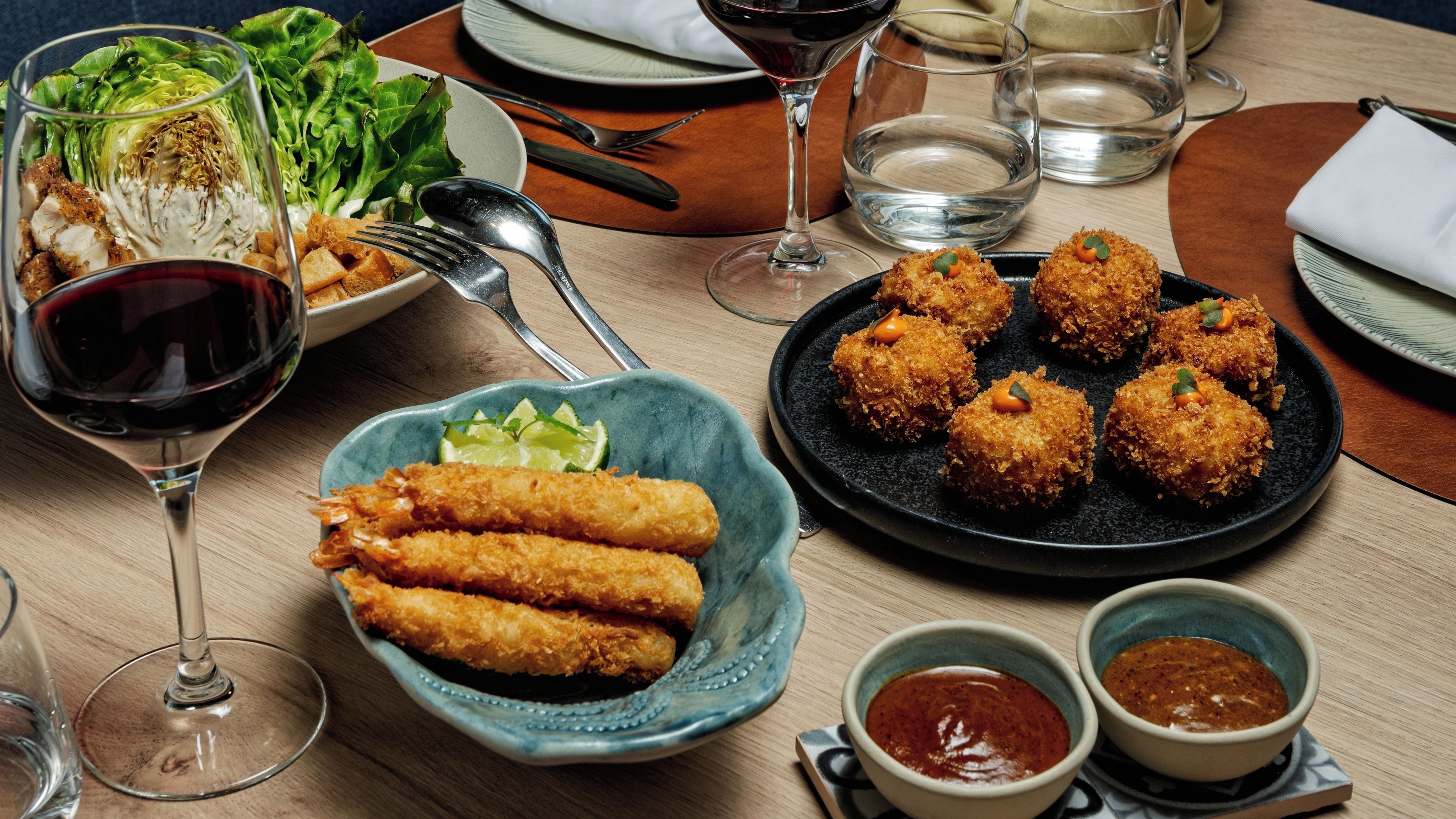 Una mesa dispuesta con ensalada, alimentos rebozados, copas de vino y pequeños recipientes de salsa.