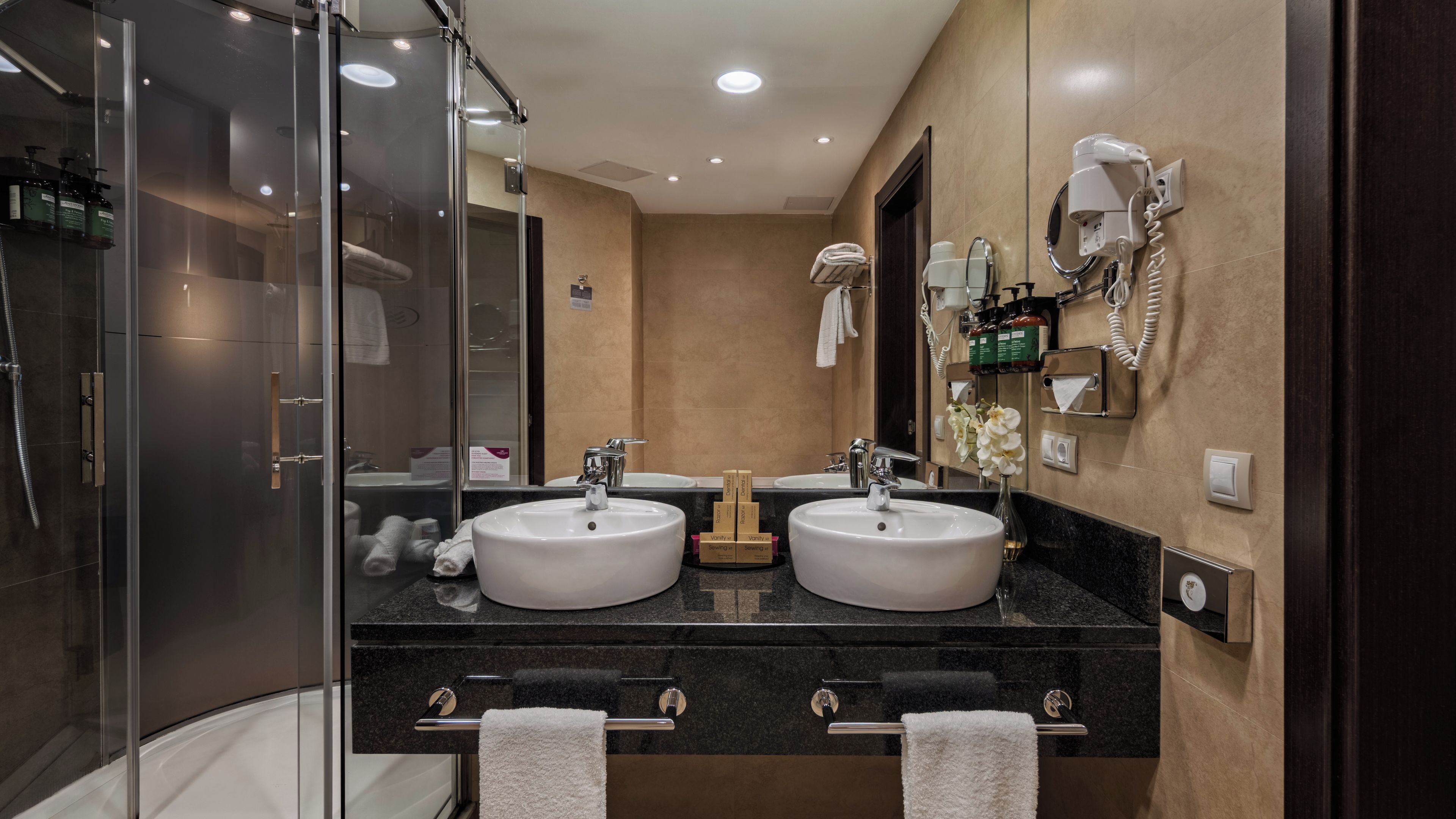 Een moderne badkamer met twee wastafels, een douchecabine van glas en een aan de muur bevestigde haardroger.