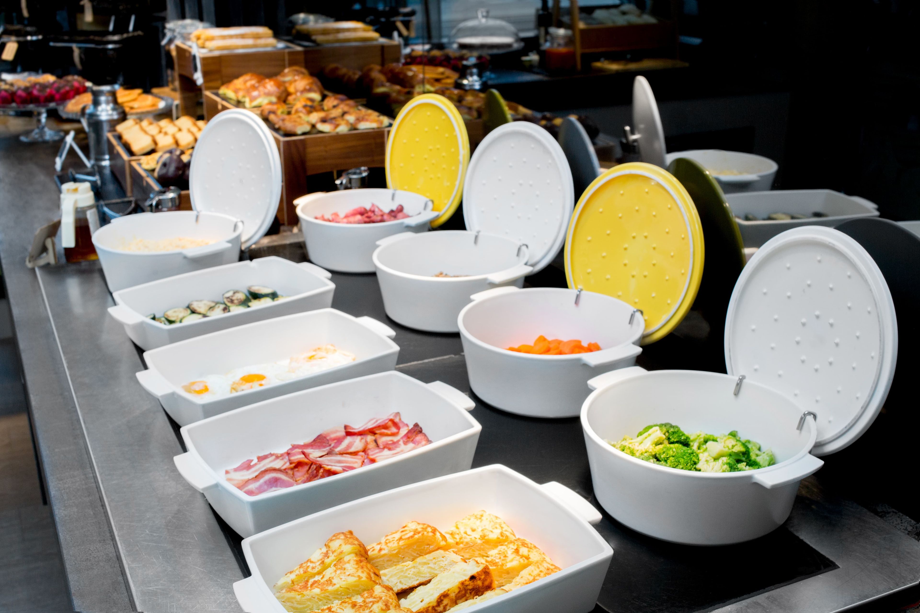 A buffet with various food items in white containers, including eggs, bacon, and vegetables.
