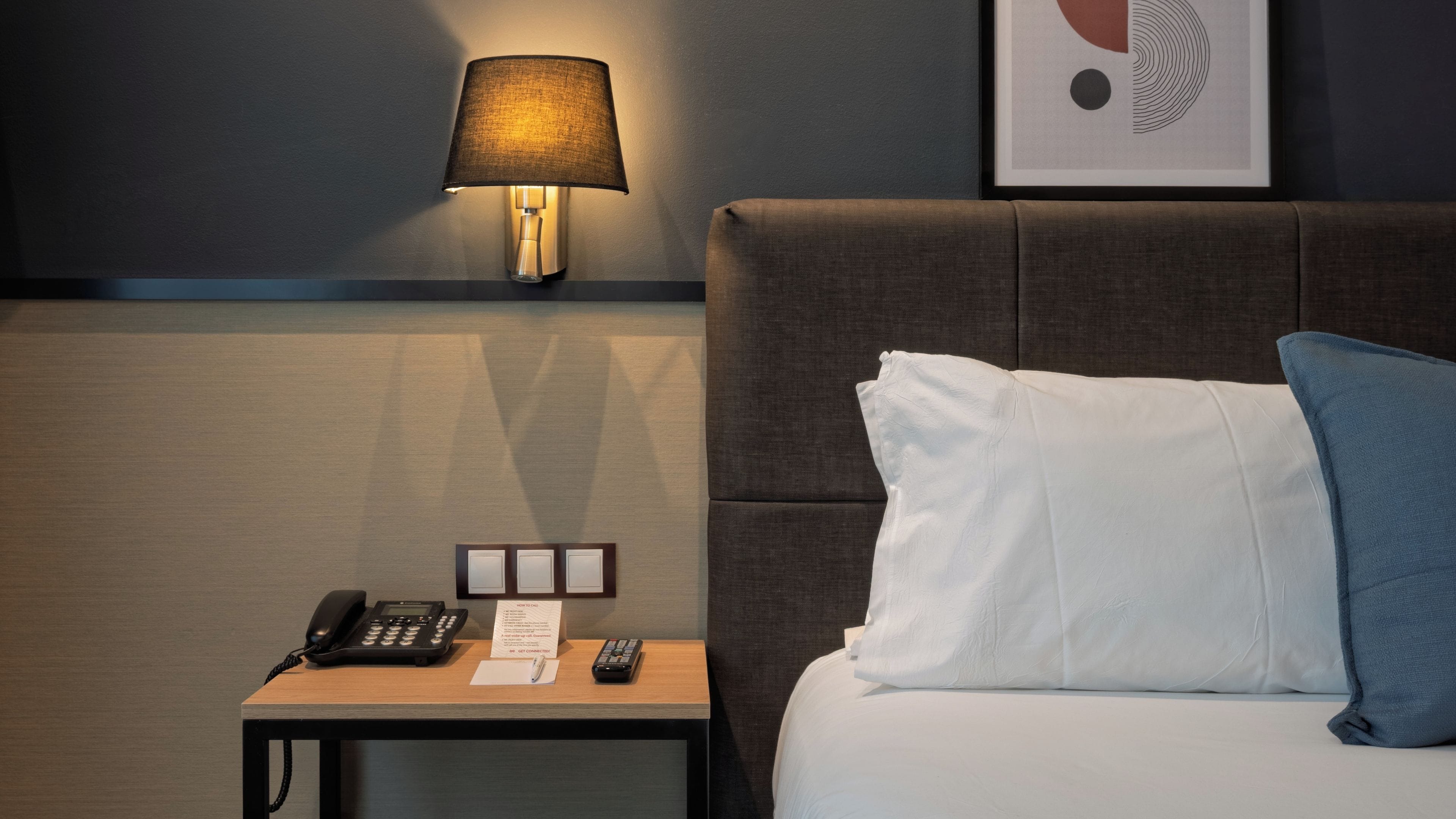 Ein Hotelzimmer mit einem Bett, einem Nachttisch, einer Lampe, einem Telefon und einem gerahmten Bild an der Wand.