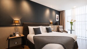Habitació d'hotel amb un llit gran, làmpades de nit, una cadira, una taula petita i un vas amb flors.