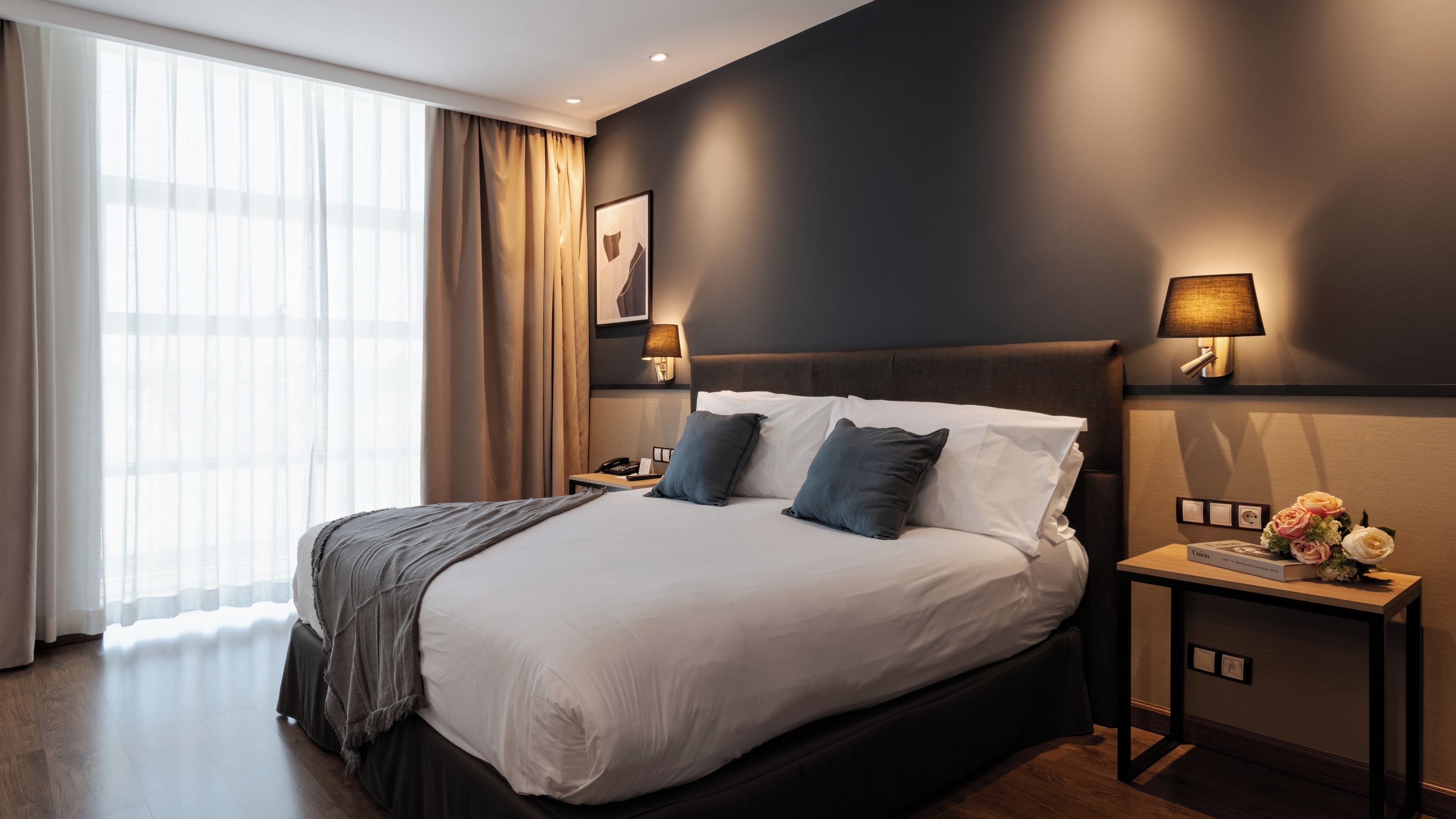 Ein Hotelzimmer mit einem großen Bett, zwei Nachttischlampen, einem Nachttisch mit Telefon und Lampe sowie einem Fenster mit Vorhängen.