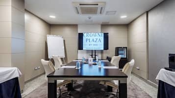 Ein Konferenzraum mit einem großen Bildschirm, der 'CROWNE PLAZA BY IHG Madrid Airport' anzeigt, einer Tafel, einem Kühlschrank und einem Tisch mit Stühlen.
