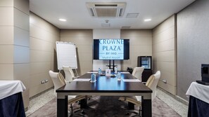 Una sala de conferències amb una pantalla gran que mostra 'CROWNE PLAZA BY IHG Madrid Airport', una pissarra, un frigorífic i una taula amb cadires.