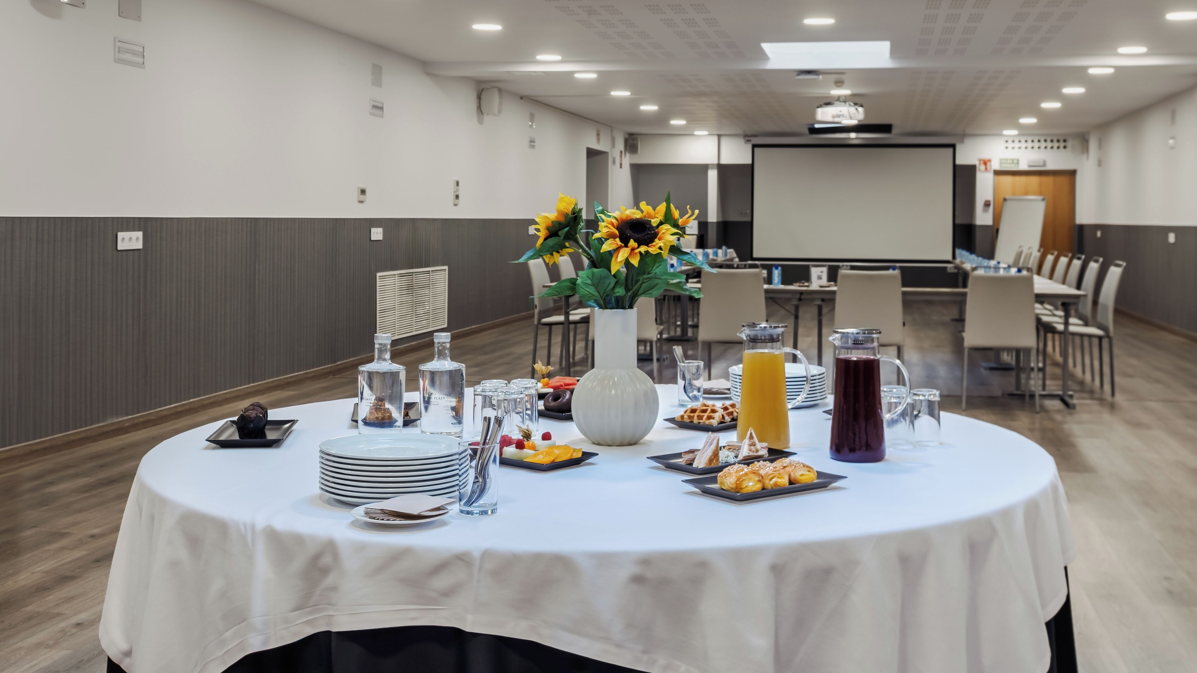 Ein Konferenzraum mit einem weißen Tisch, der für eine Besprechung gedeckt ist, ausgestattet mit einer Projektionsleinwand und umgeben von Stühlen.