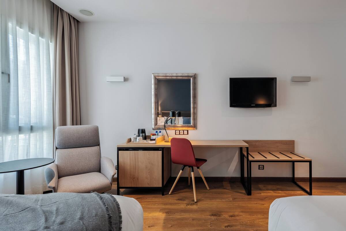 Una habitación de hotel con cama, escritorio, silla, televisión y ventana con cortinas.