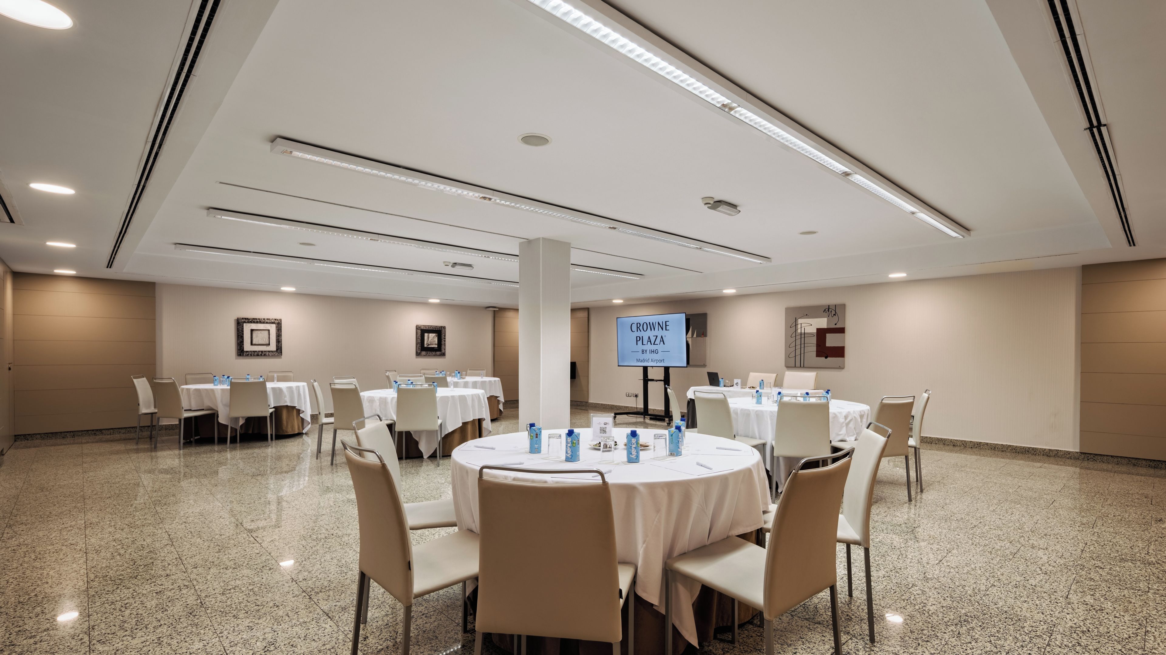 Un salón de conferencias con mesas redondas, sillas y una gran pantalla que muestra 'CROWNE PLAZA BY IHG Madrid Airport'.