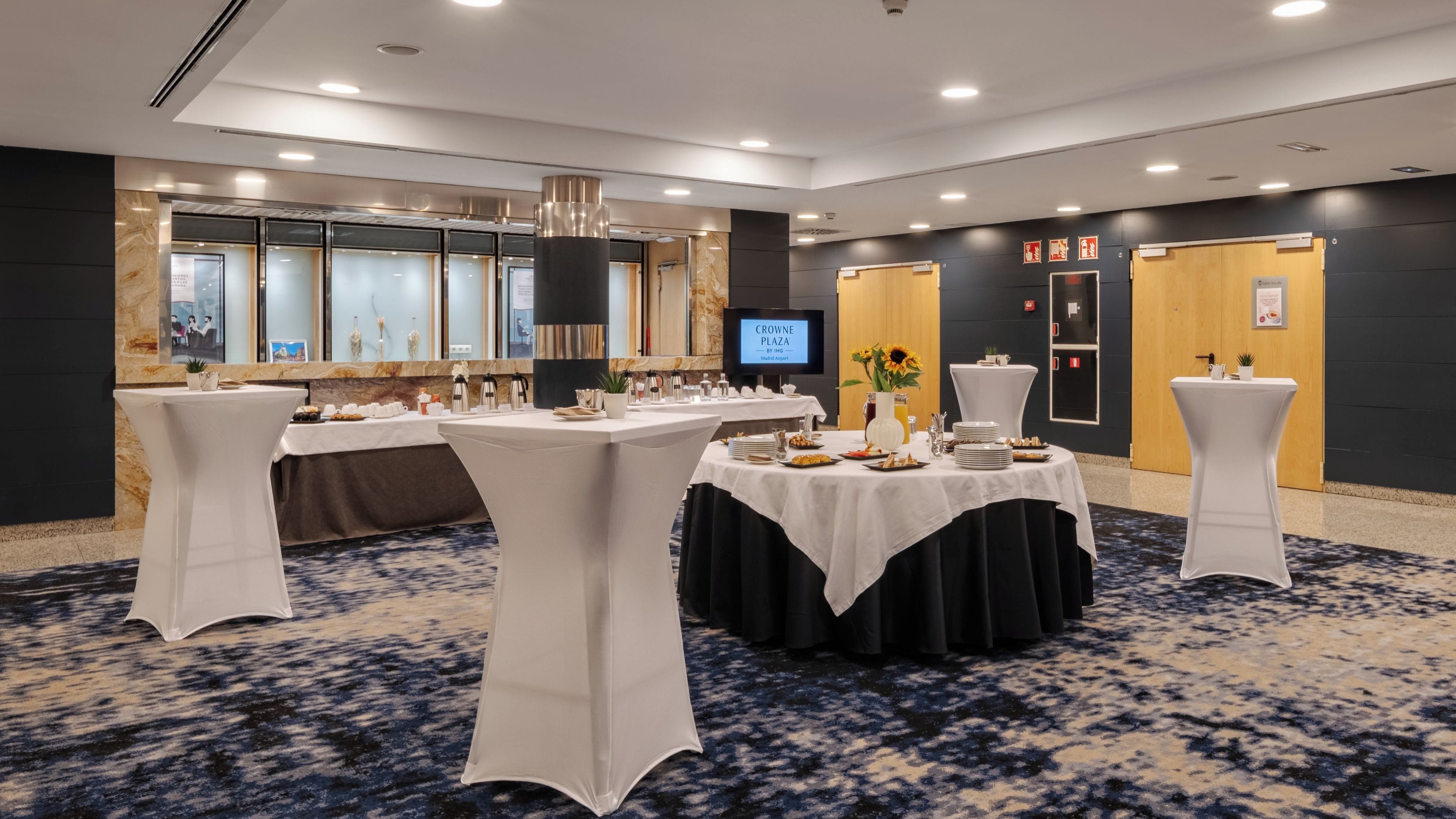 A conference room with round tables, white tablecloths, and a large screen displaying 'CROWNE PLAZA BY IHG Madrid Airport'.