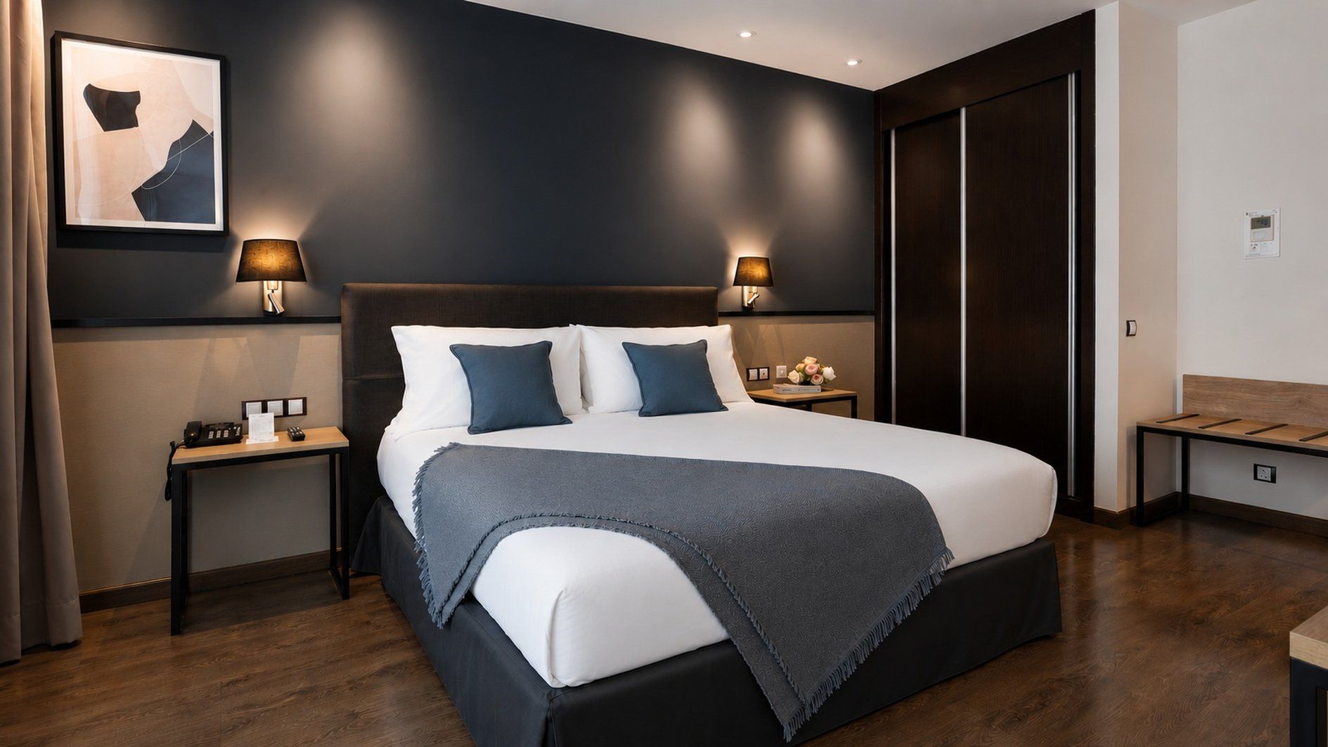 Habitación de hotel moderna con una cama grande, dos mesitas de noche, un cabecero y una pared oscura con una obra de arte enmarcada.
