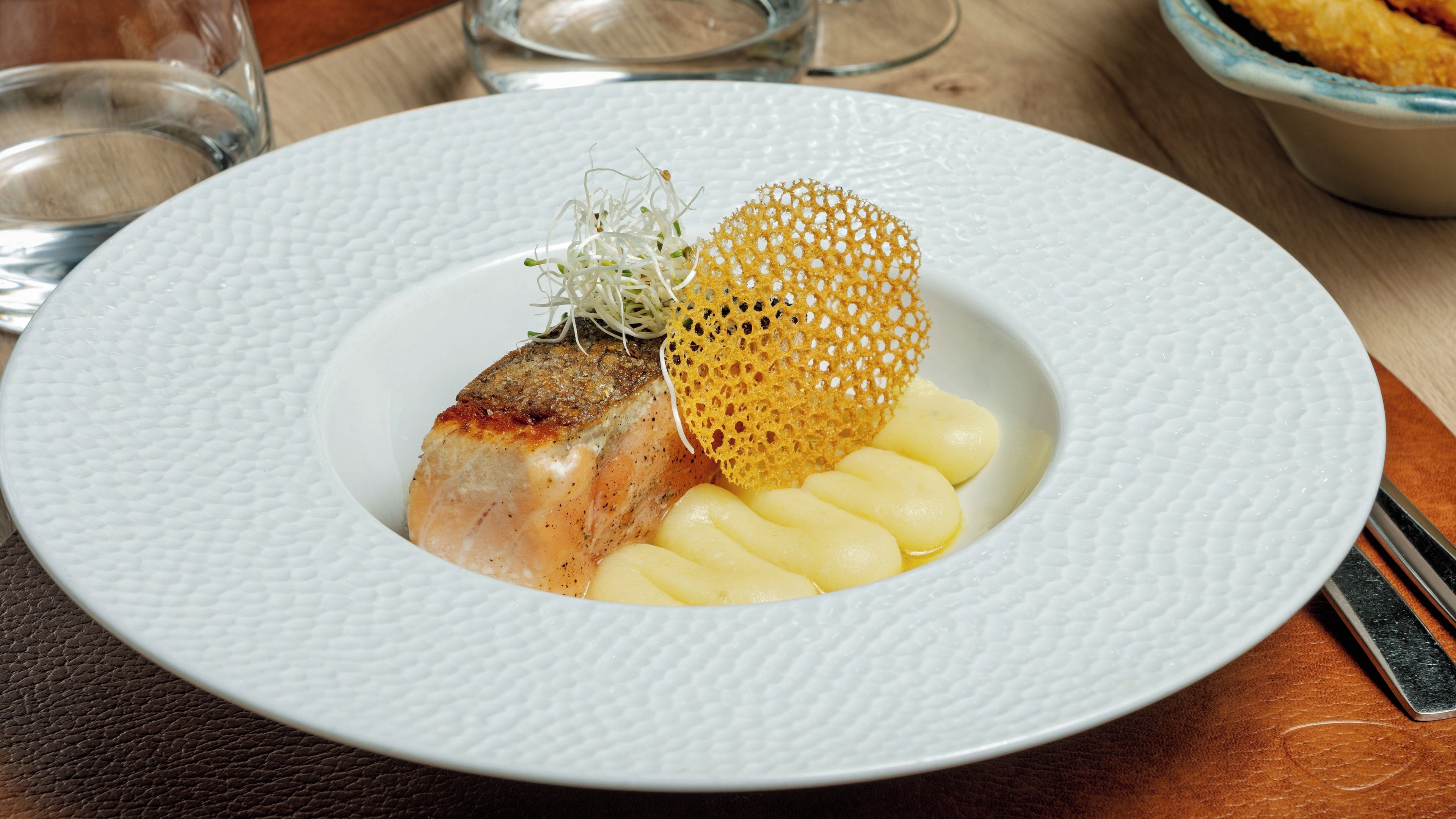 A white textured plate with a piece of fish, a yellowish garnish, and some white elements on a wooden table.