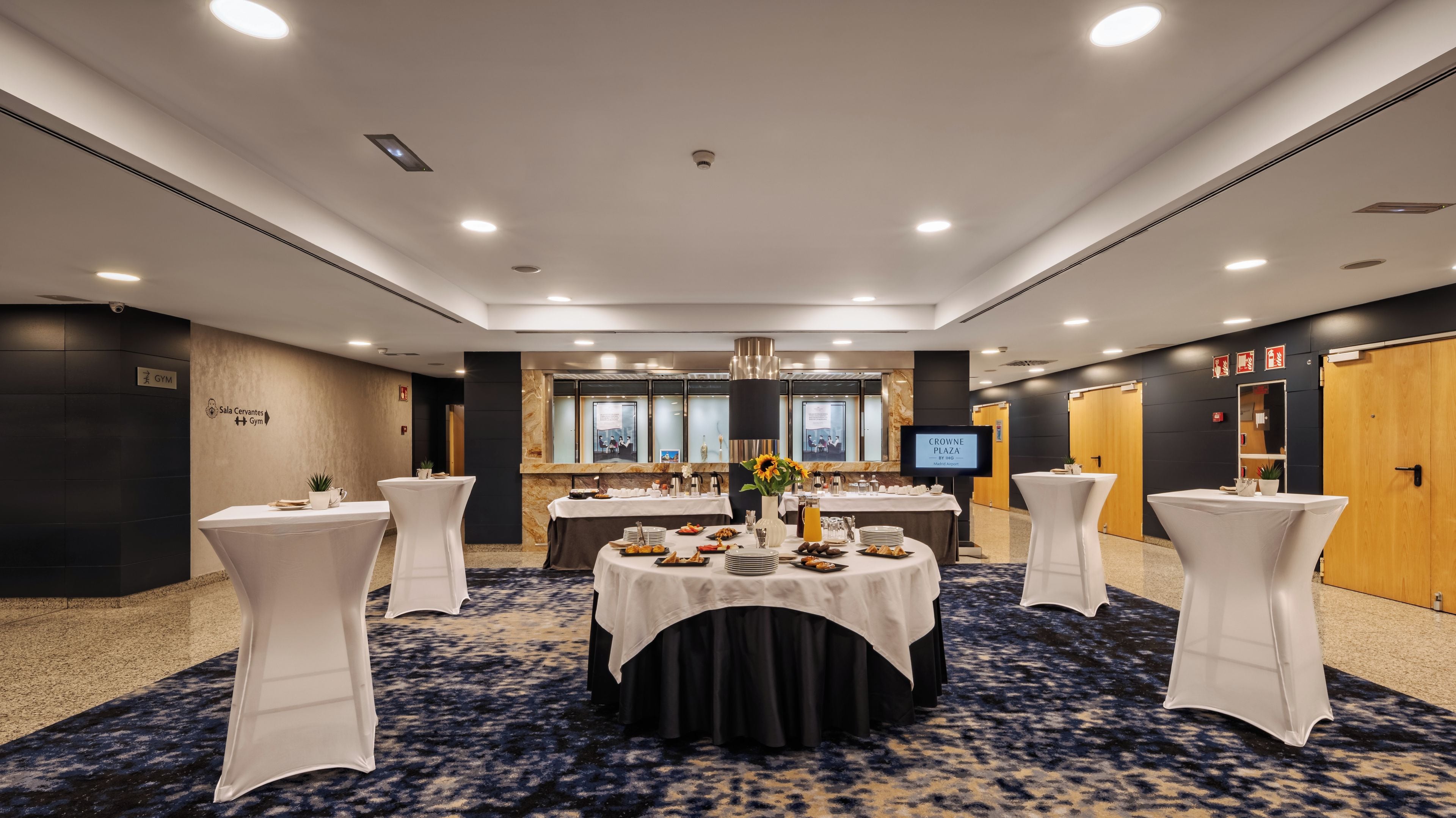 Un salón de conferencias con mesas redondas, sillas blancas, una pantalla grande y un suelo cubierto de alfombra oscura.