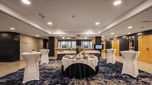 A conference room with round tables, white chairs, a large screen, and a dark carpeted floor. - Crowne Plaza Madrid Airport by IHG (Madrid)